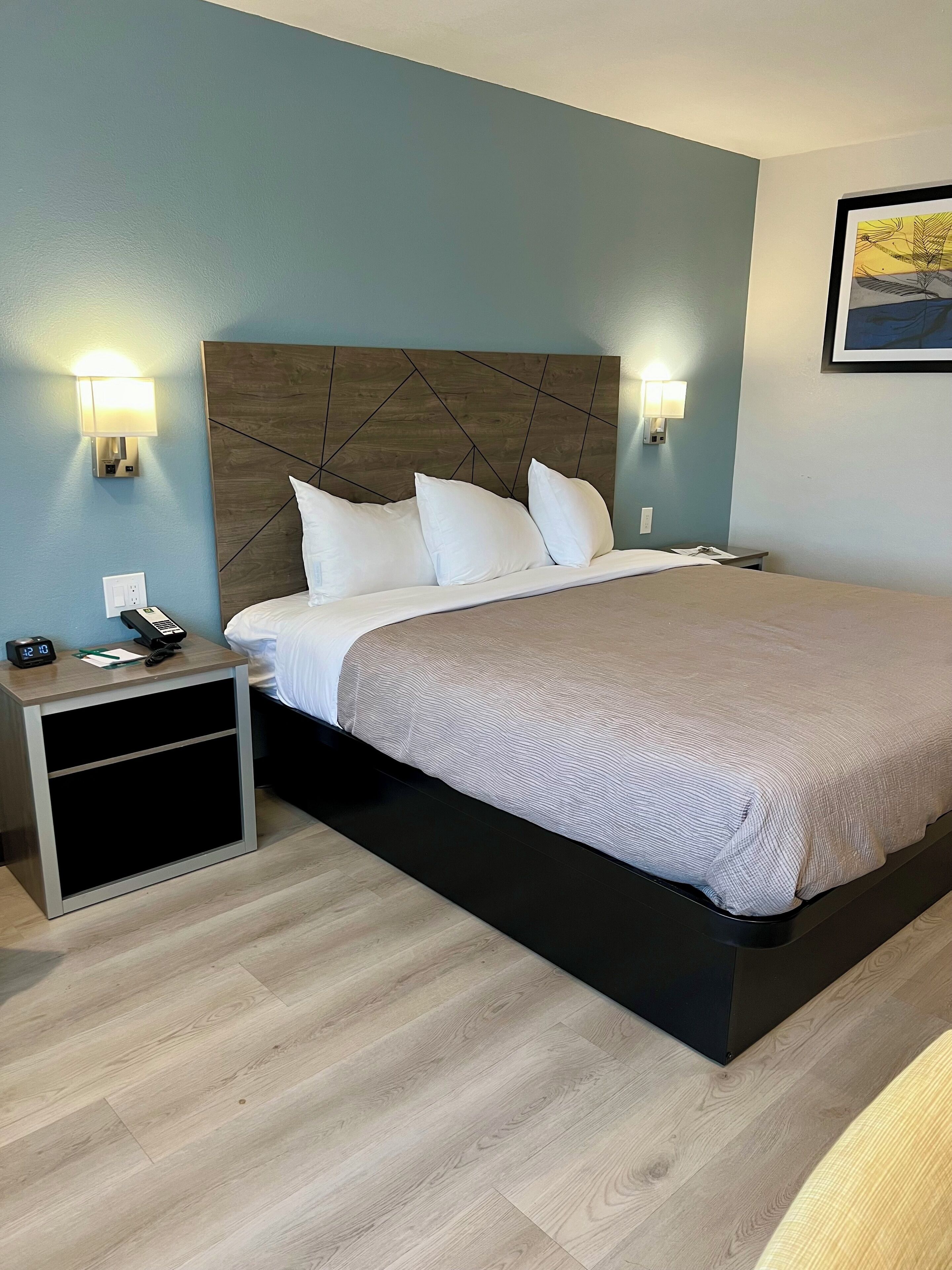 A modern hotel room with a wooden headboard, a large bed with white linens, a bedside table with a phone and a clock, and wall-mounted lights.
