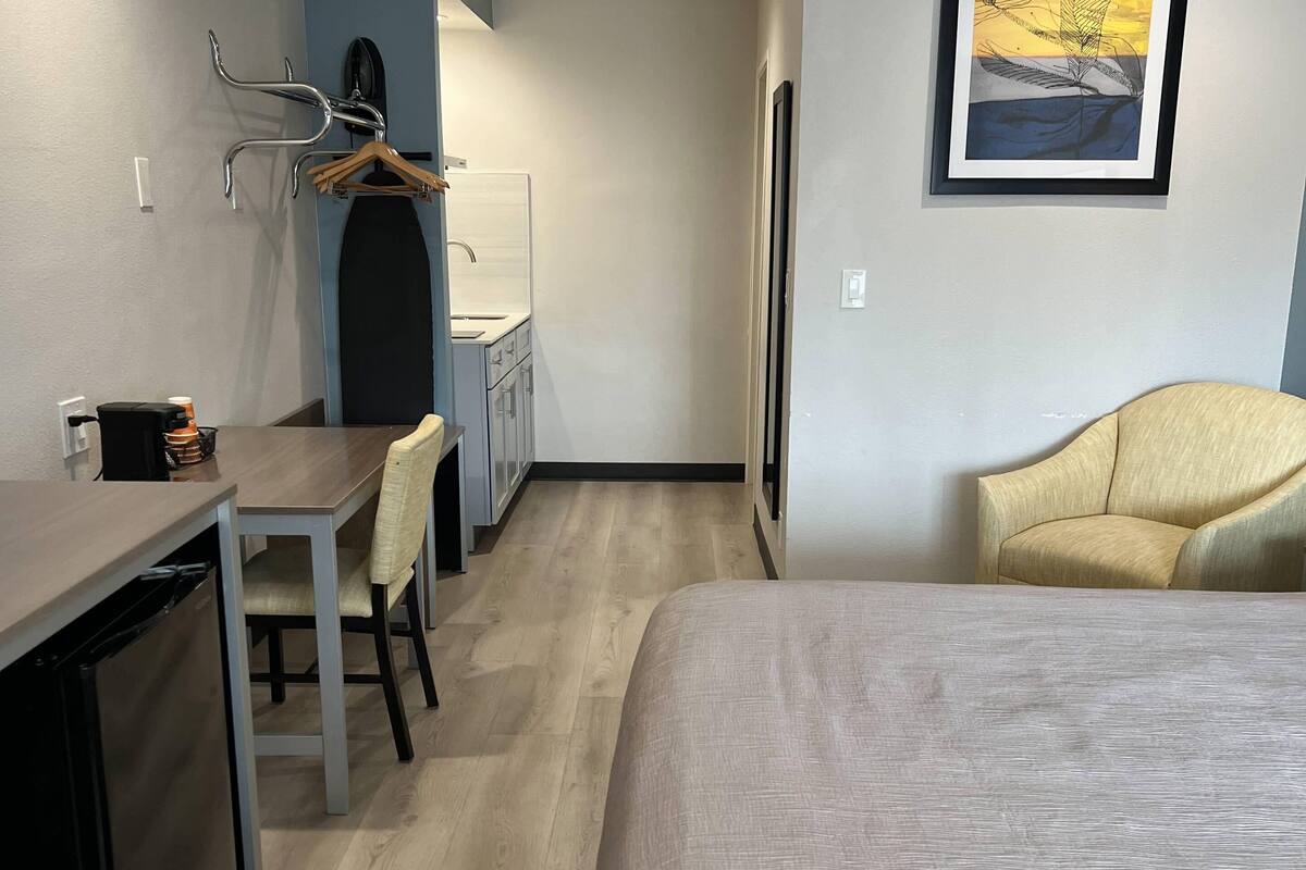 A hotel room with a bed, a desk with a chair, a small kitchenette, and a chair with a cushion.