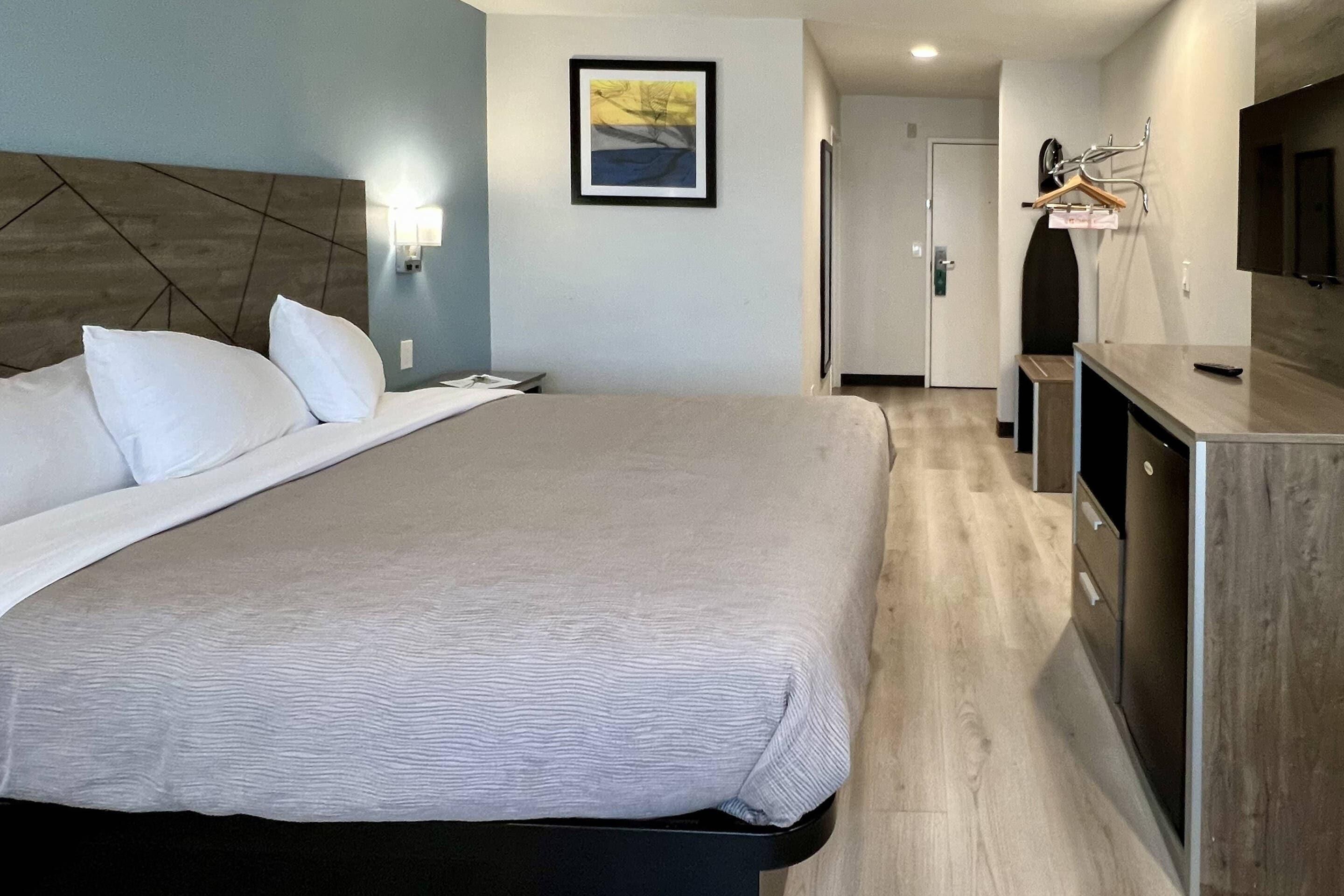 Une chambre d’hôtel avec un grand lit, une tête de lit en bois, un téléviseur fixé au mur, une table de chevet et une porte donnant sur un couloir.