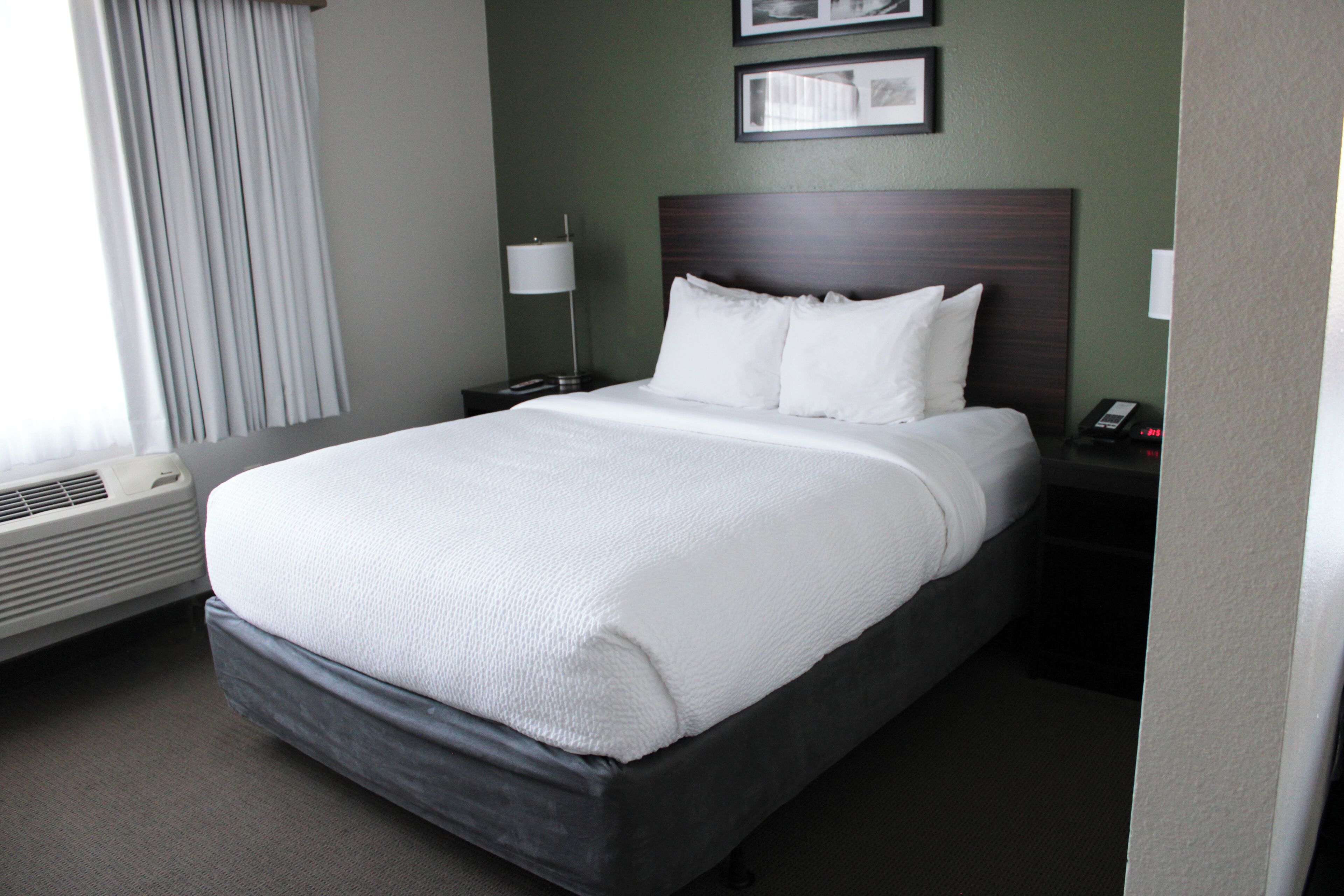 A neatly made bed with white linens in a hotel room with green walls, a dark headboard, and a bedside table with a lamp.