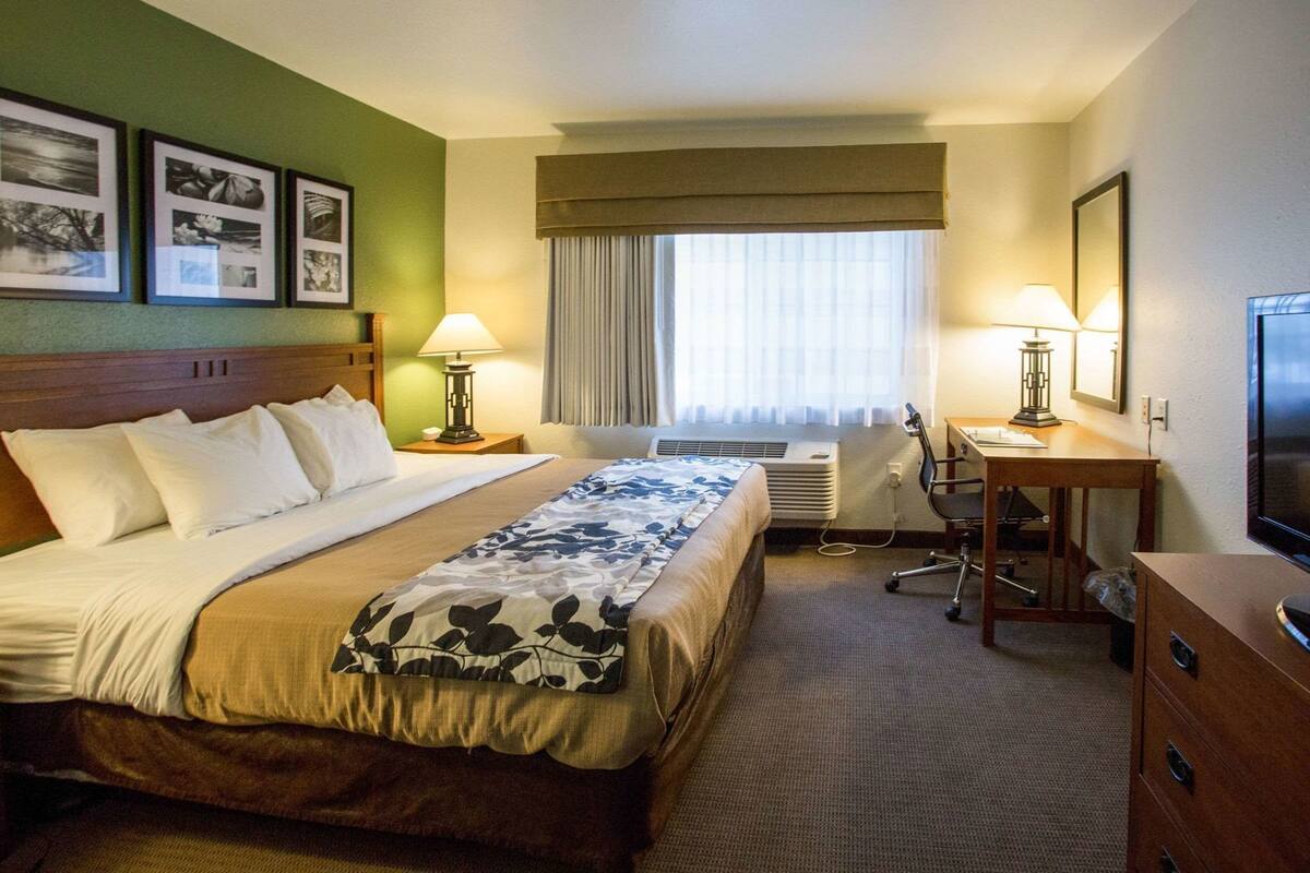 A hotel room with a bed, desk, chair, television, air conditioning unit, and framed pictures on the wall.