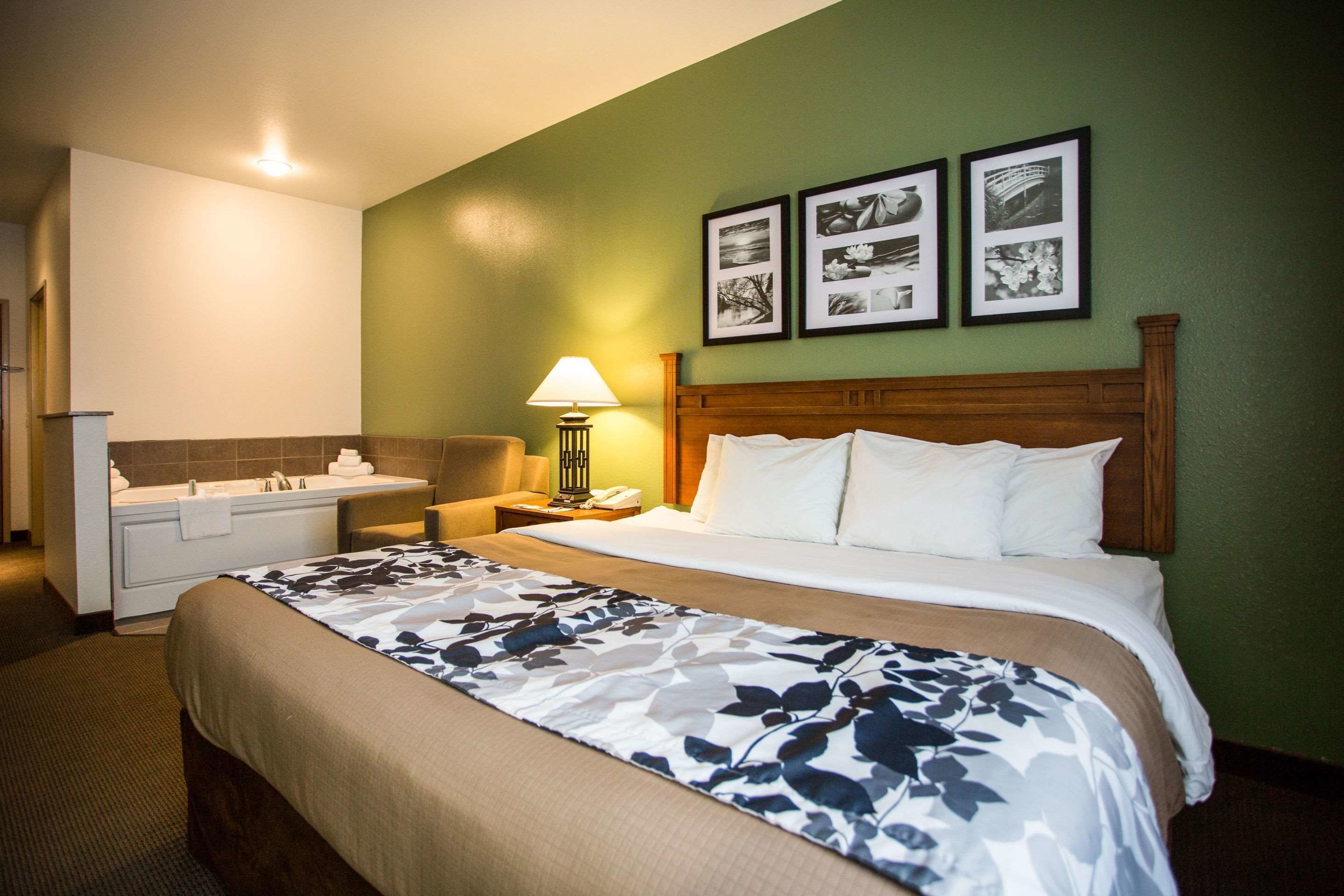 A hotel room with a bed, a nightstand, a lamp, a bathroom with a bathtub, and a wall with framed pictures.