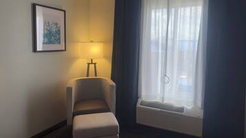 Habitación de hotel con una silla, una lámpara, un cuadro en la pared y una ventana con cortinas.