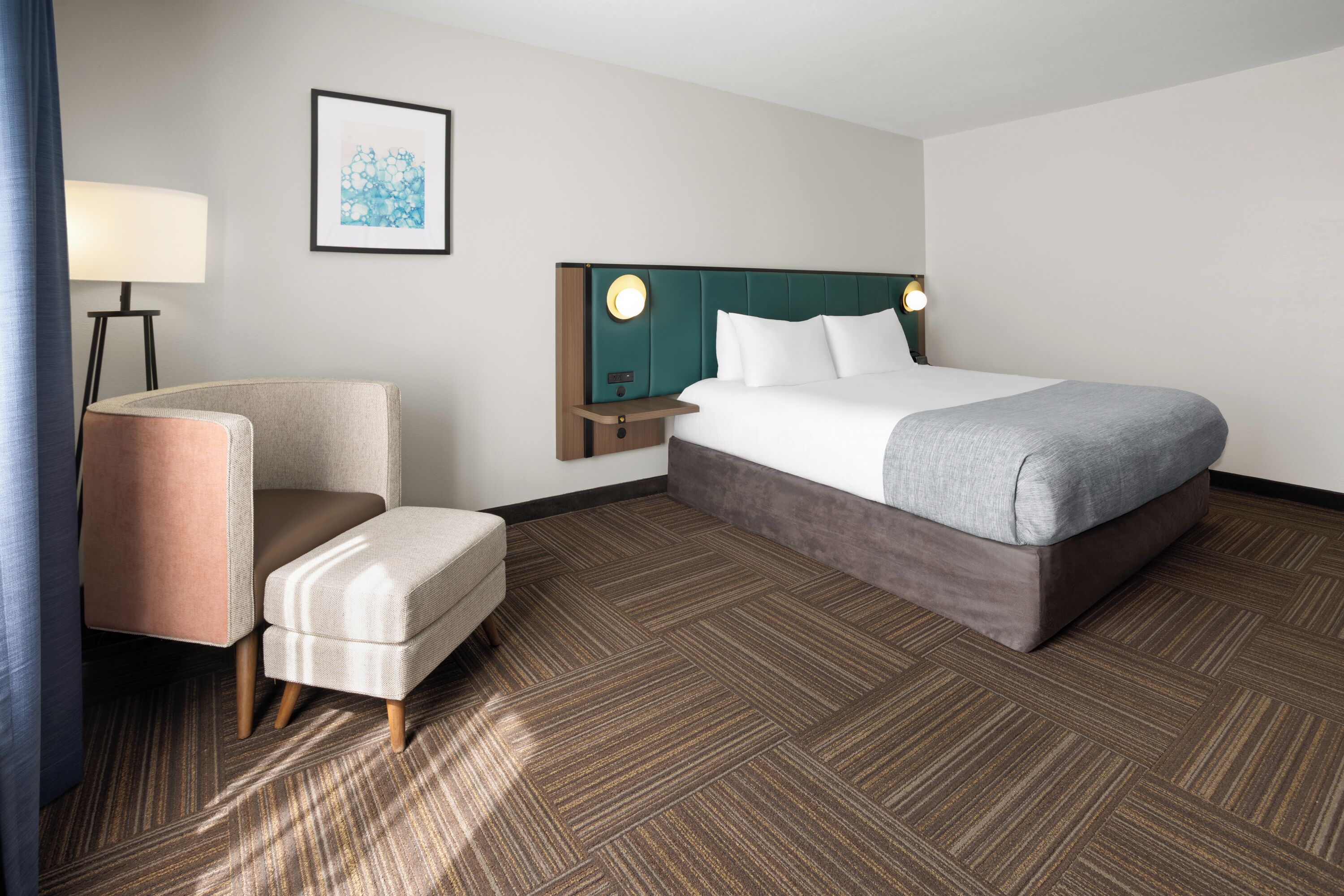 Habitación de hotel con cama, sillón, mesita de noche, lámpara y un cuadro en la pared.