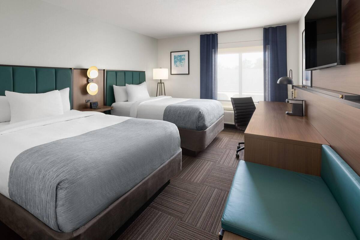 Ein Hotelzimmer mit zwei Betten, einem Schreibtisch, einem Stuhl, einem Fernseher und einem Fenster mit Vorhängen.