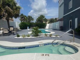 Una piscina con indicador de profundidad de 90 centímetros, rodeada de sillas de descanso y palmeras, con un edificio al fondo.