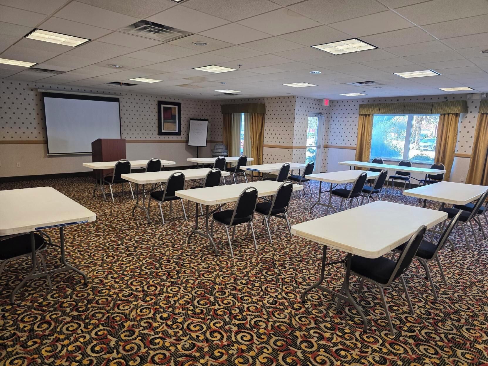 A conference room with white tables and chairs, a projector screen, and a patterned carpet.