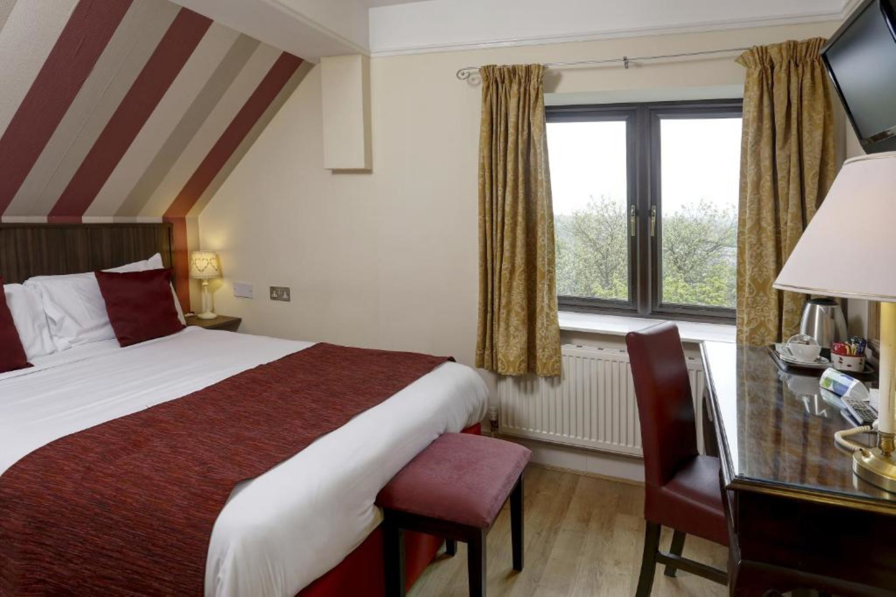 A hotel room with a large bed, a desk with a lamp, a chair, and a window with curtains.