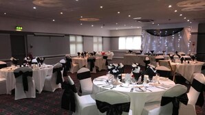 Un salón de banquetes con mesas redondas preparadas para un evento formal, cada una con mantel blanco y fajín negro.