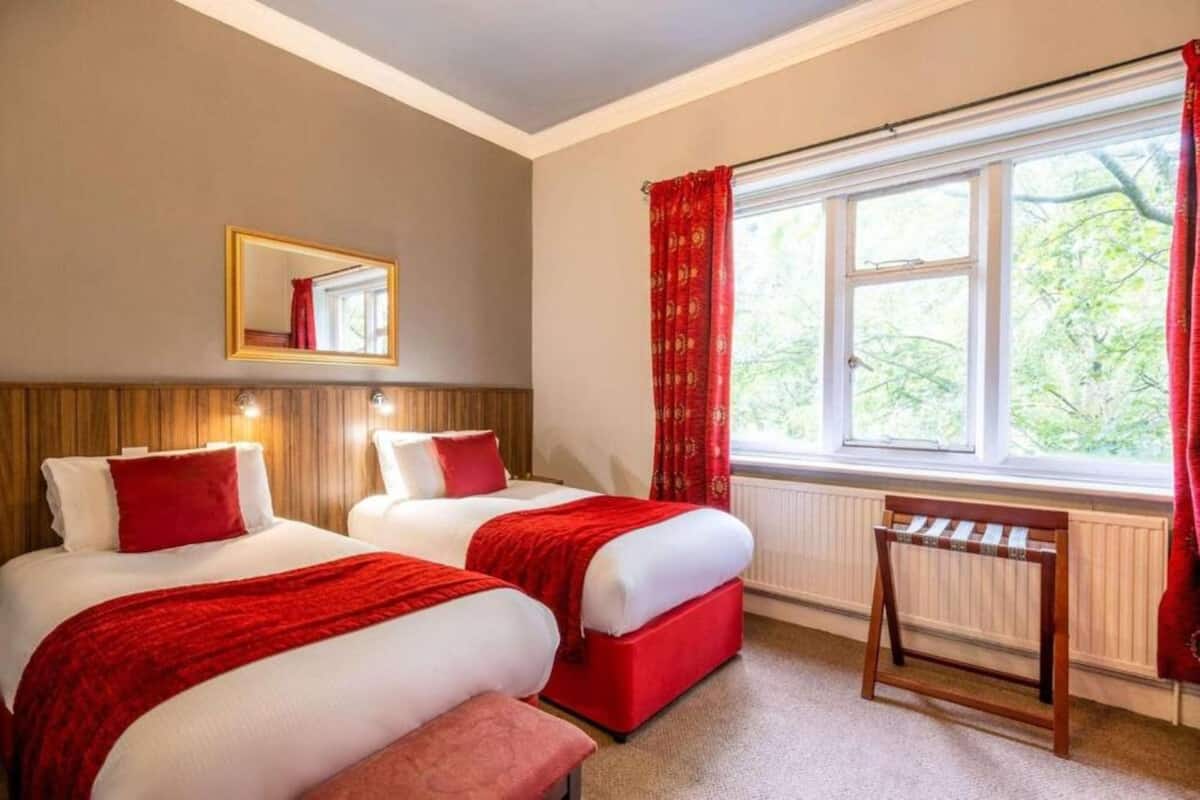 Una camera d'albergo con due letti, un termosifone, una finestra con tende rosse e uno specchio appeso al muro.