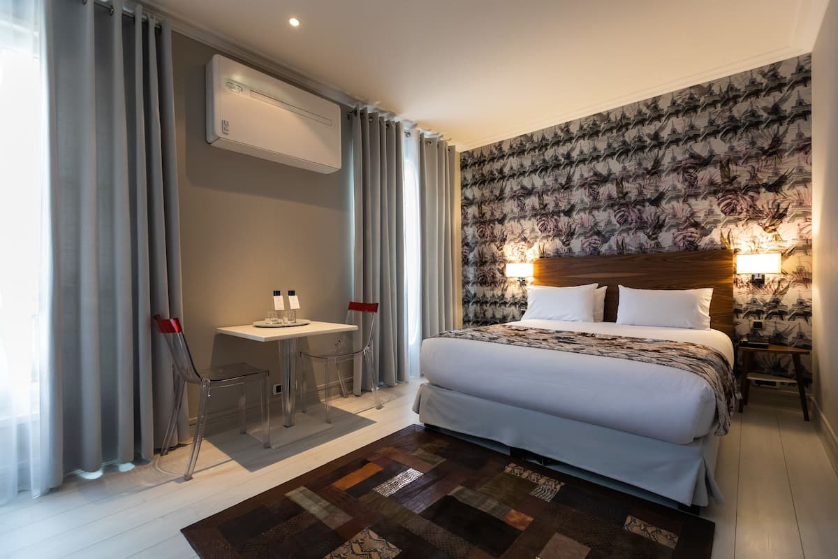 A hotel room with a large bed, a desk with two chairs, an air conditioning unit, and a patterned wallpaper.
