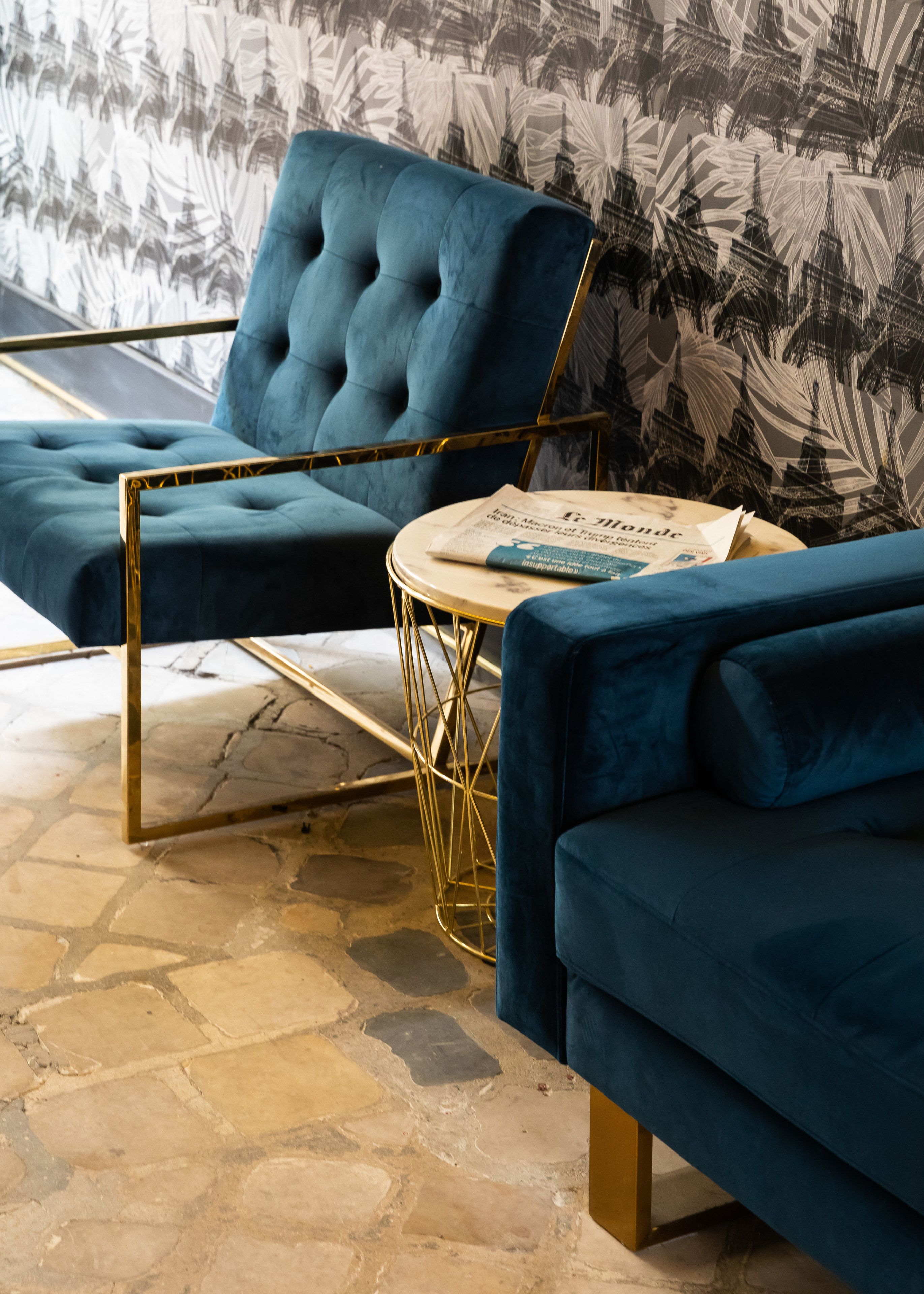 A blue armchair with a gold frame, a round coffee table, and a blue sofa in a room with a patterned wallpaper and stone flooring.
