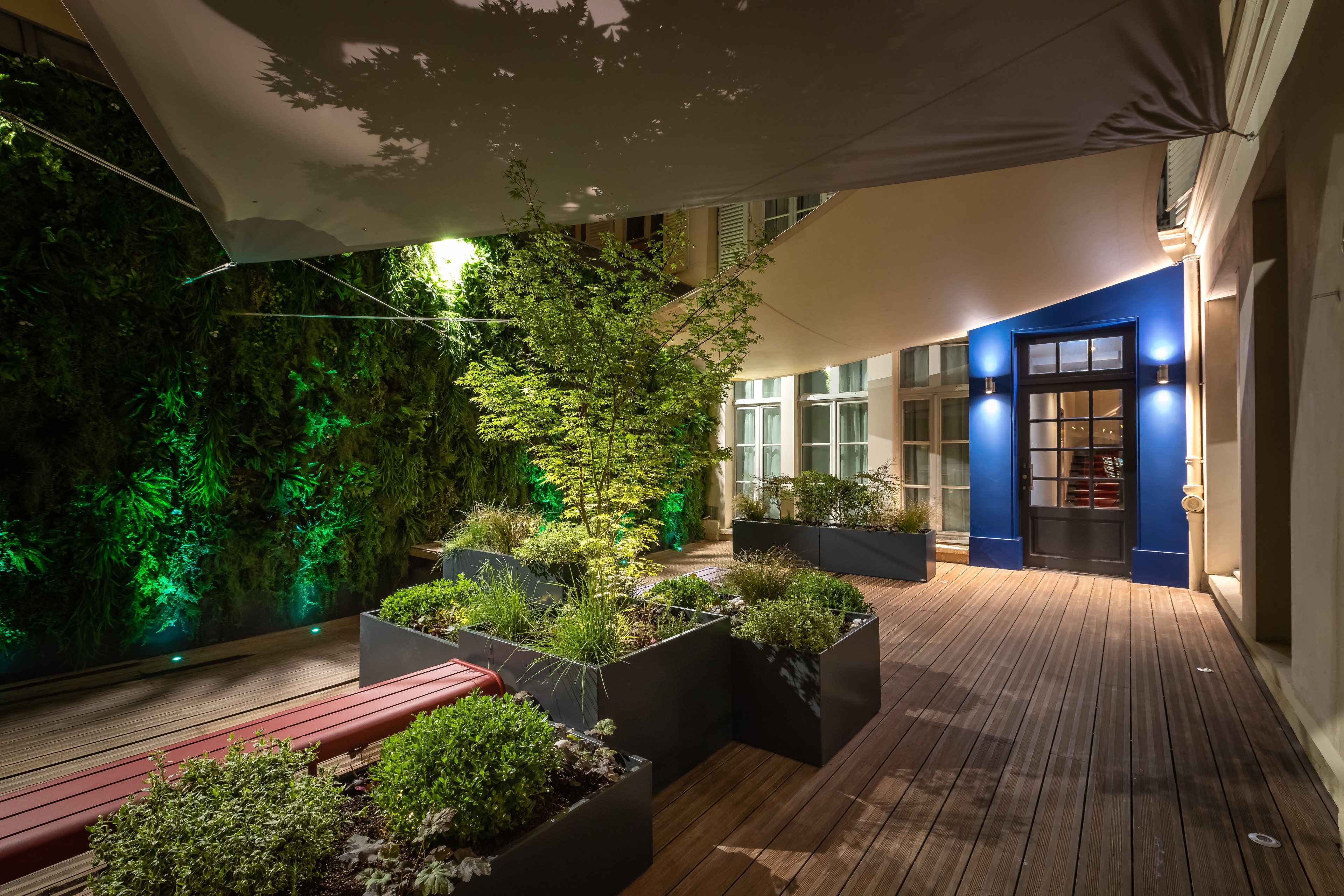 A modern outdoor patio with wooden flooring, black planters with greenery, and a blue wall with a door.