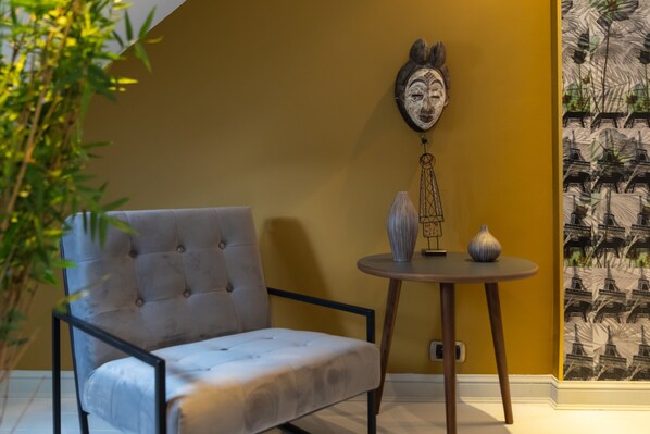 Una habitación con un sillón tapizado, una pequeña mesa redonda con adornos, y una pared con papel tapiz estampado.