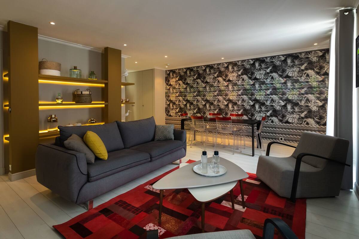 A modern living room with a grey sofa, a coffee table, and a red patterned rug. There is a wall with a leaf pattern and a TV mounted on the wall.