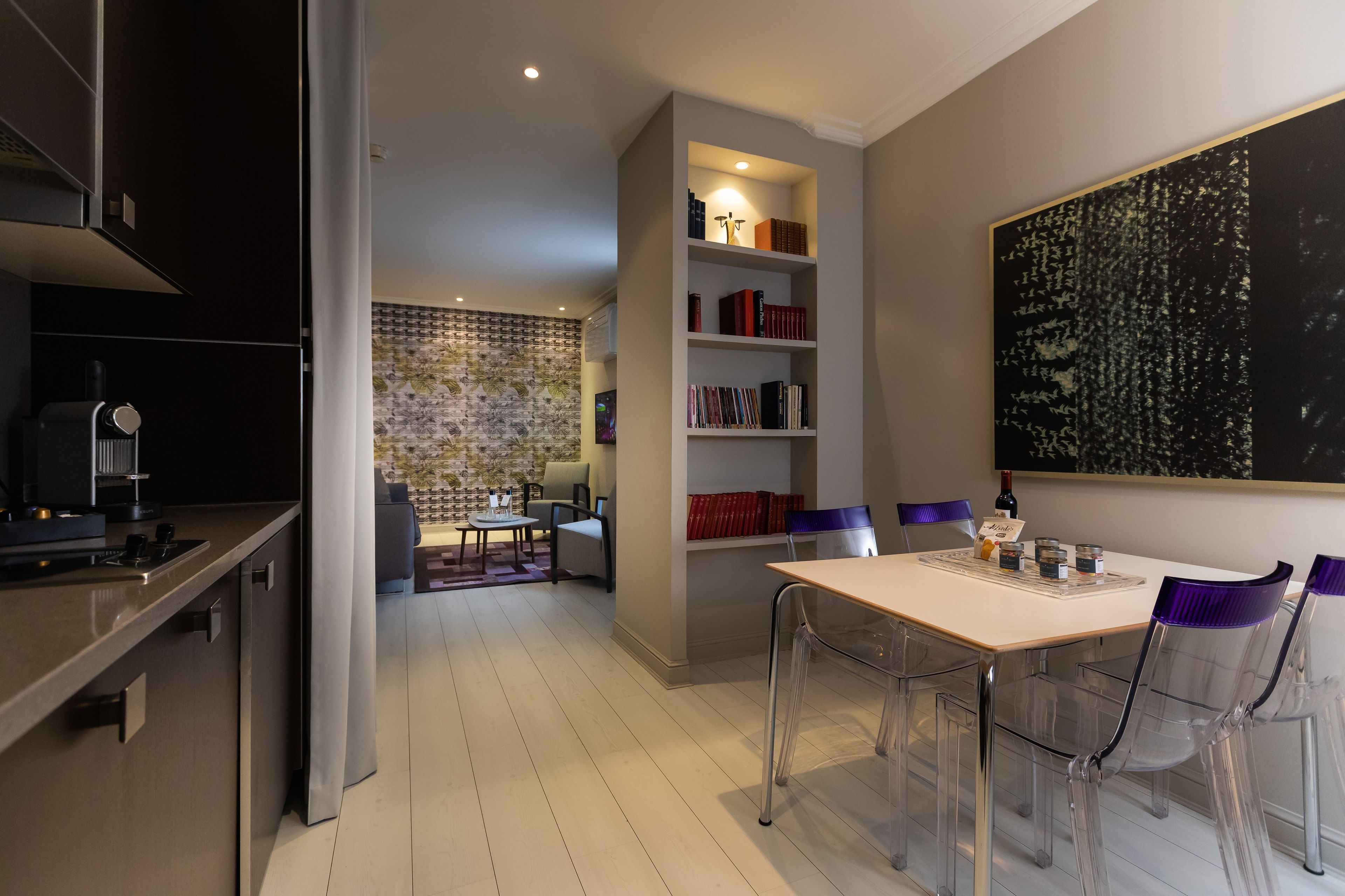 A modern kitchen with a built-in oven, a dining area with a white table and clear chairs, and a bookshelf with books and decorative items.