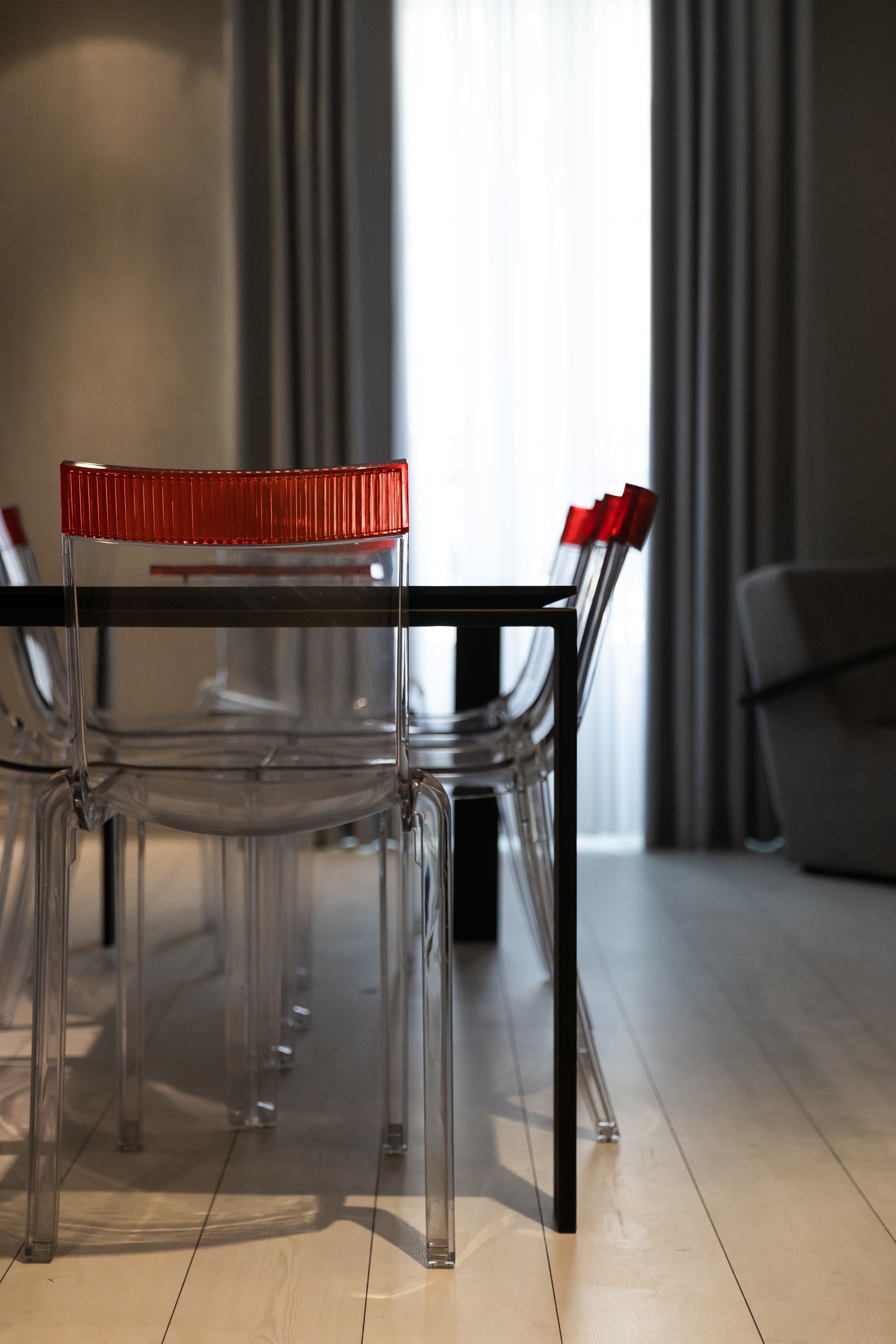 Una mesa de comedor de cristal transparente con detalles en rojo, rodeada de sillas transparentes, en una habitación moderna con suelo de madera y un sofá gris al fondo.