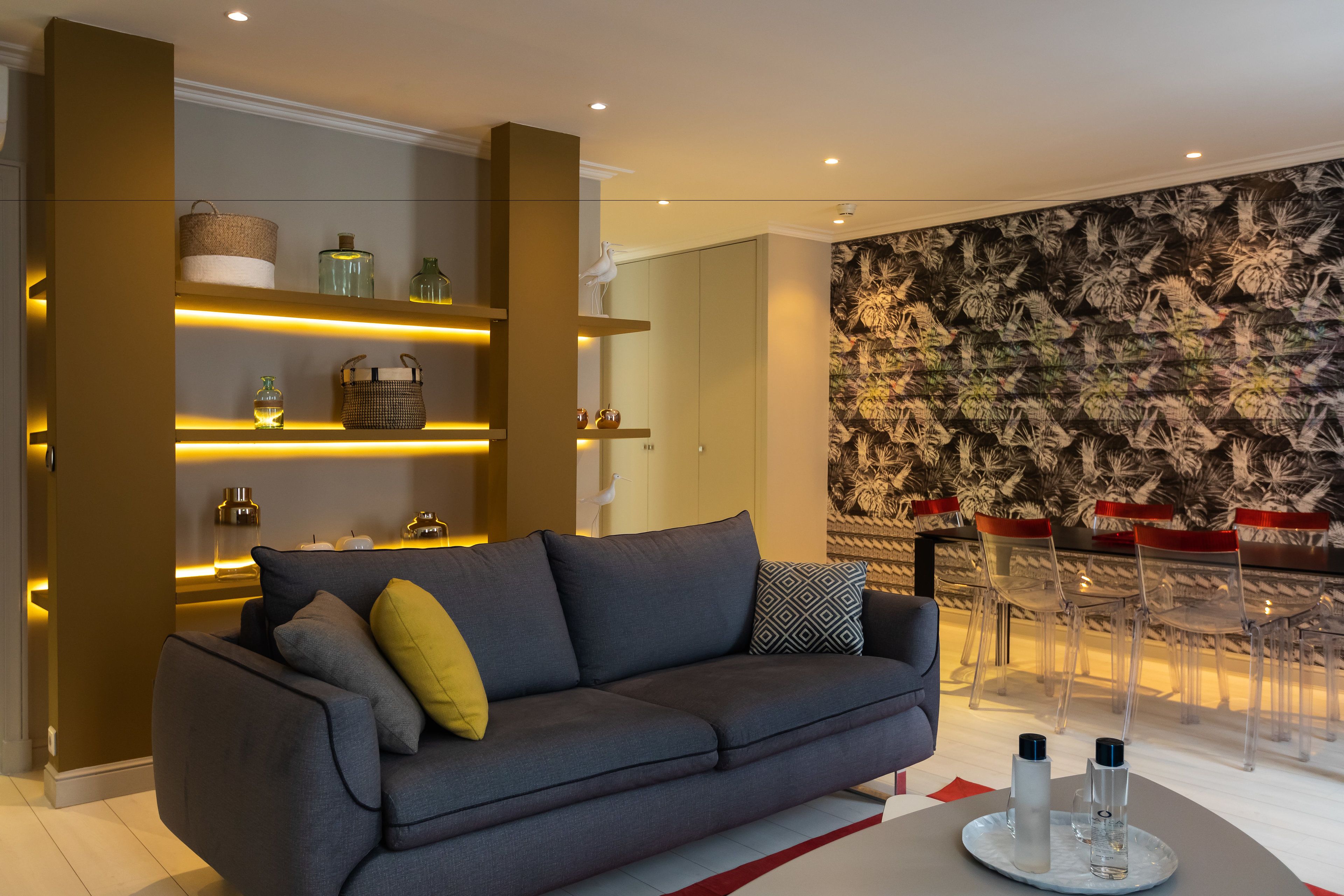 A modern living room with a grey sofa, shelves with decorative items, a patterned wall, and a dining area with clear chairs.
