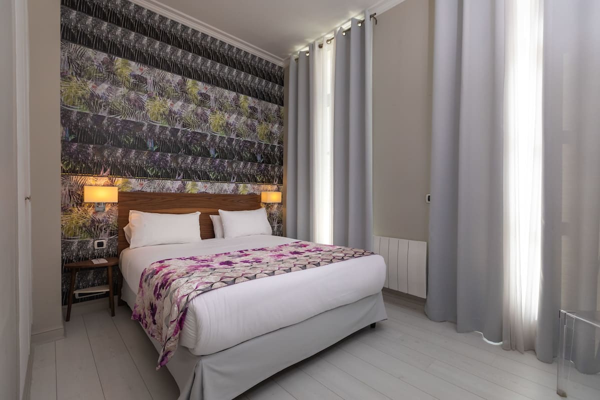 A neatly arranged bedroom with a large bed, a nightstand, a wall-mounted lamp, and a window with curtains.