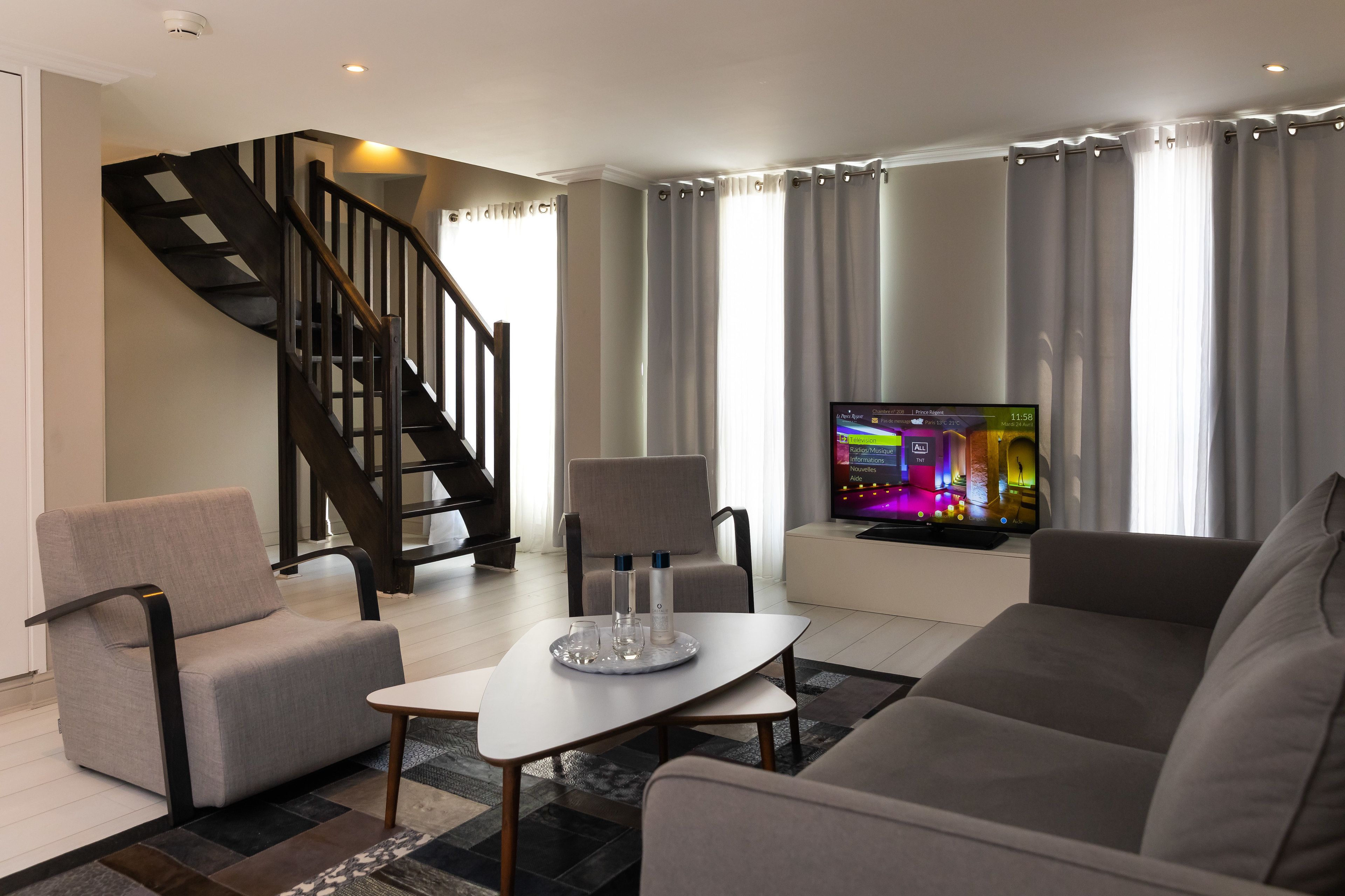 Ein modernes Wohnzimmer mit einer Couch, Sesseln, einem Couchtisch und einem Fernseher auf einem Lowboard. Im Hintergrund ist eine Treppe zu sehen.
