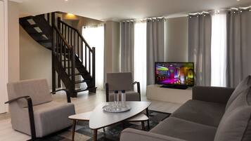 Ein modernes Wohnzimmer mit einer Couch, Sesseln, einem Couchtisch und einem Fernseher auf einem Lowboard. Im Hintergrund ist eine Treppe zu sehen.