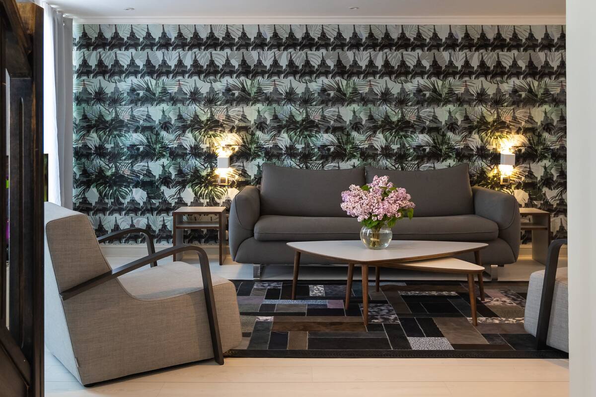 A modern living room with a grey sofa, a beige armchair, and a coffee table with a vase of flowers. The room features a patterned wallpaper and two wall-mounted lamps.