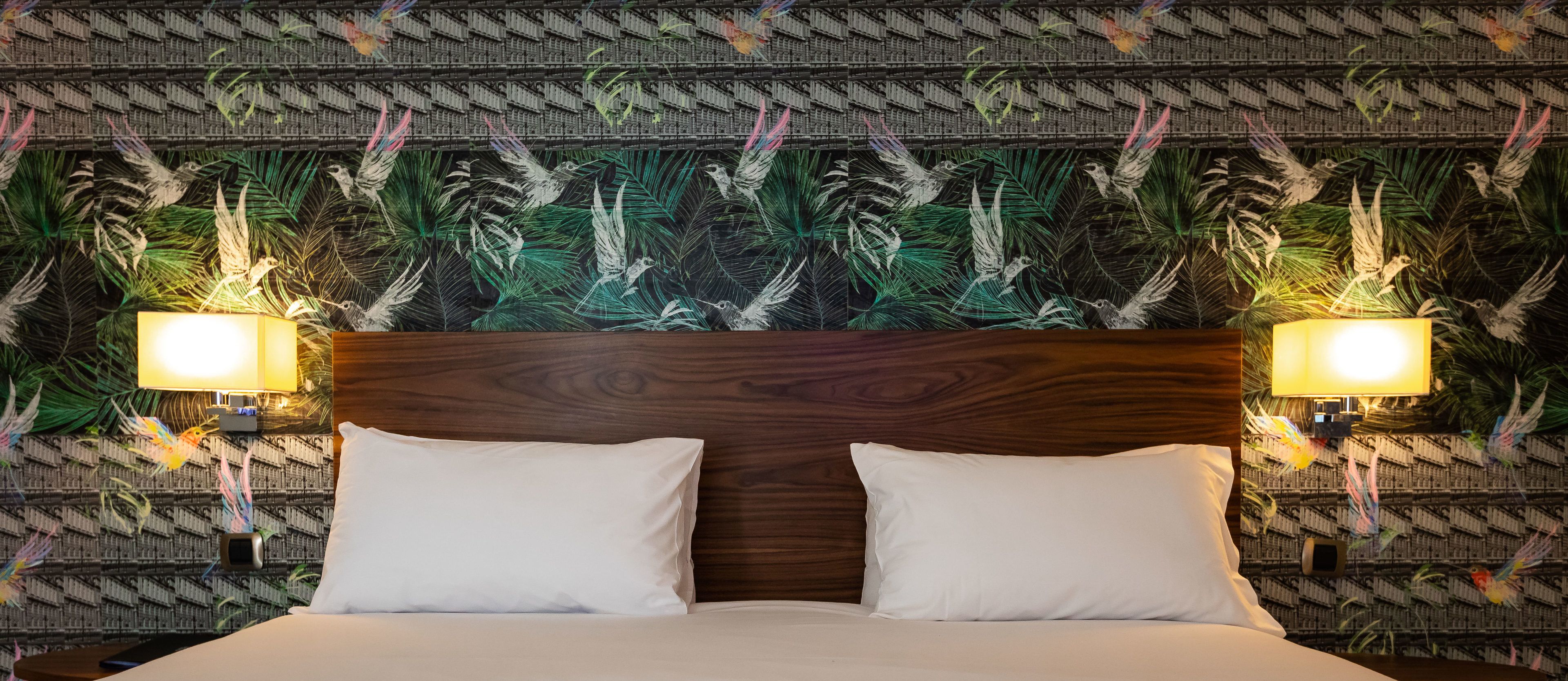 Une chambre d’hôtel avec une tête de lit en bois, deux oreillers blancs et un mur orné d’un motif de feuilles et de guirlandes lumineuses.