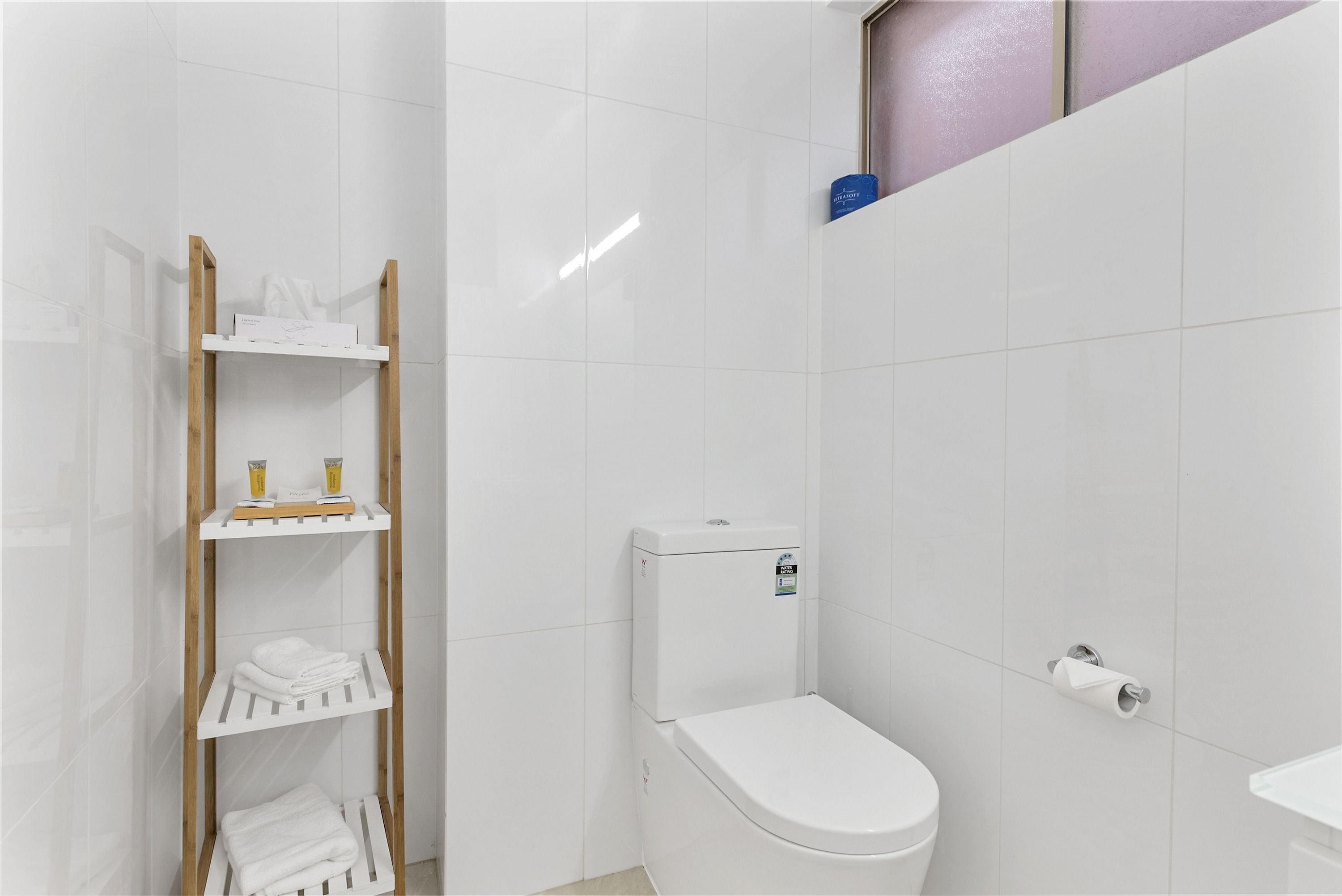 A modern bathroom with white tiles, a toilet, a shelf with toiletries, and a wooden rack holding towels.