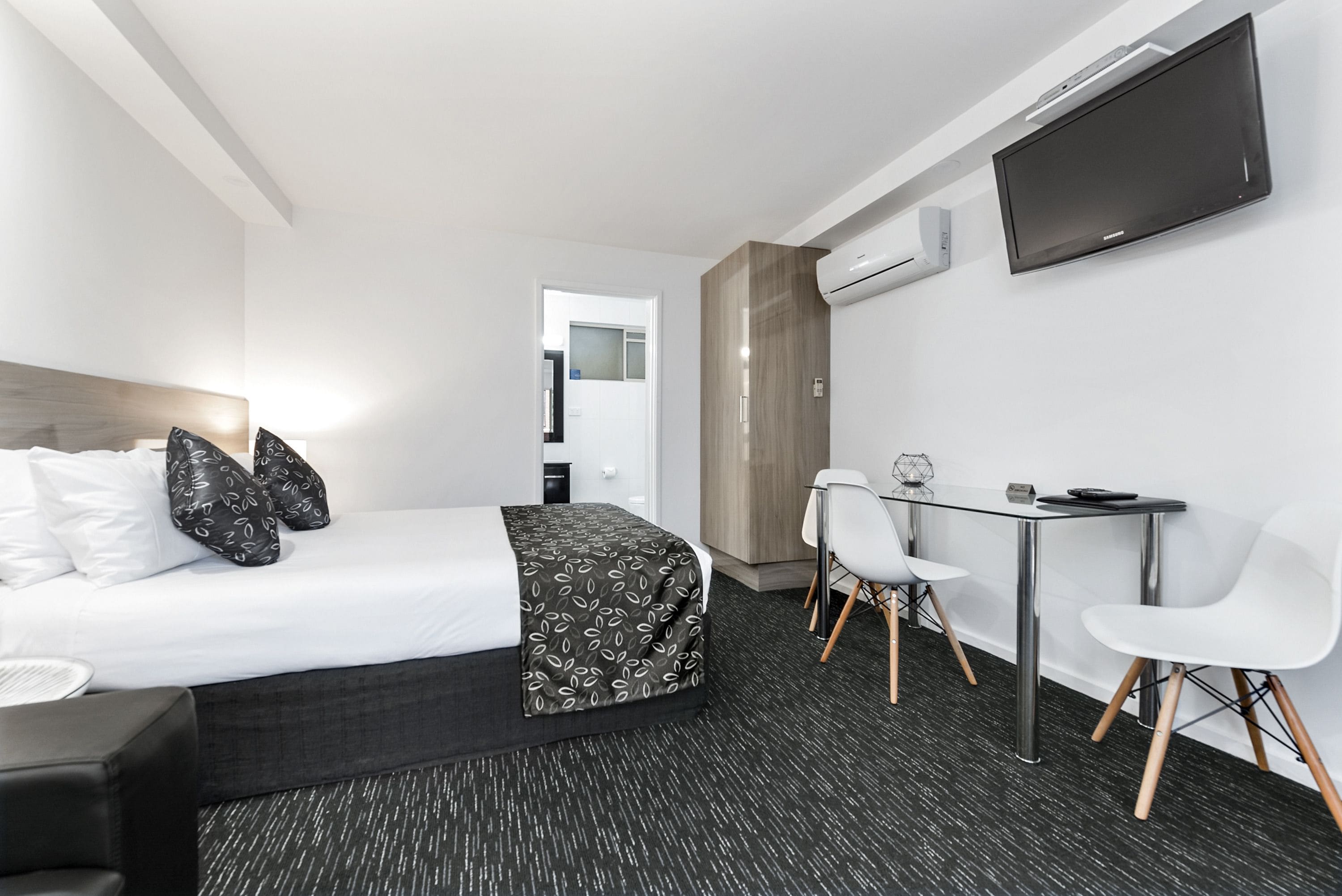 A modern hotel room with a neatly made bed, a desk with two chairs, a flat-screen TV mounted on the wall, and an air conditioning unit.
