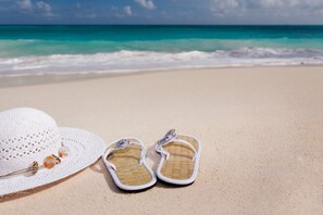 A pair of white flip-flops and a white straw hat on a sandy beach with turquoise water in the background. - Casa Giuditta Appartamenti (Palermo)