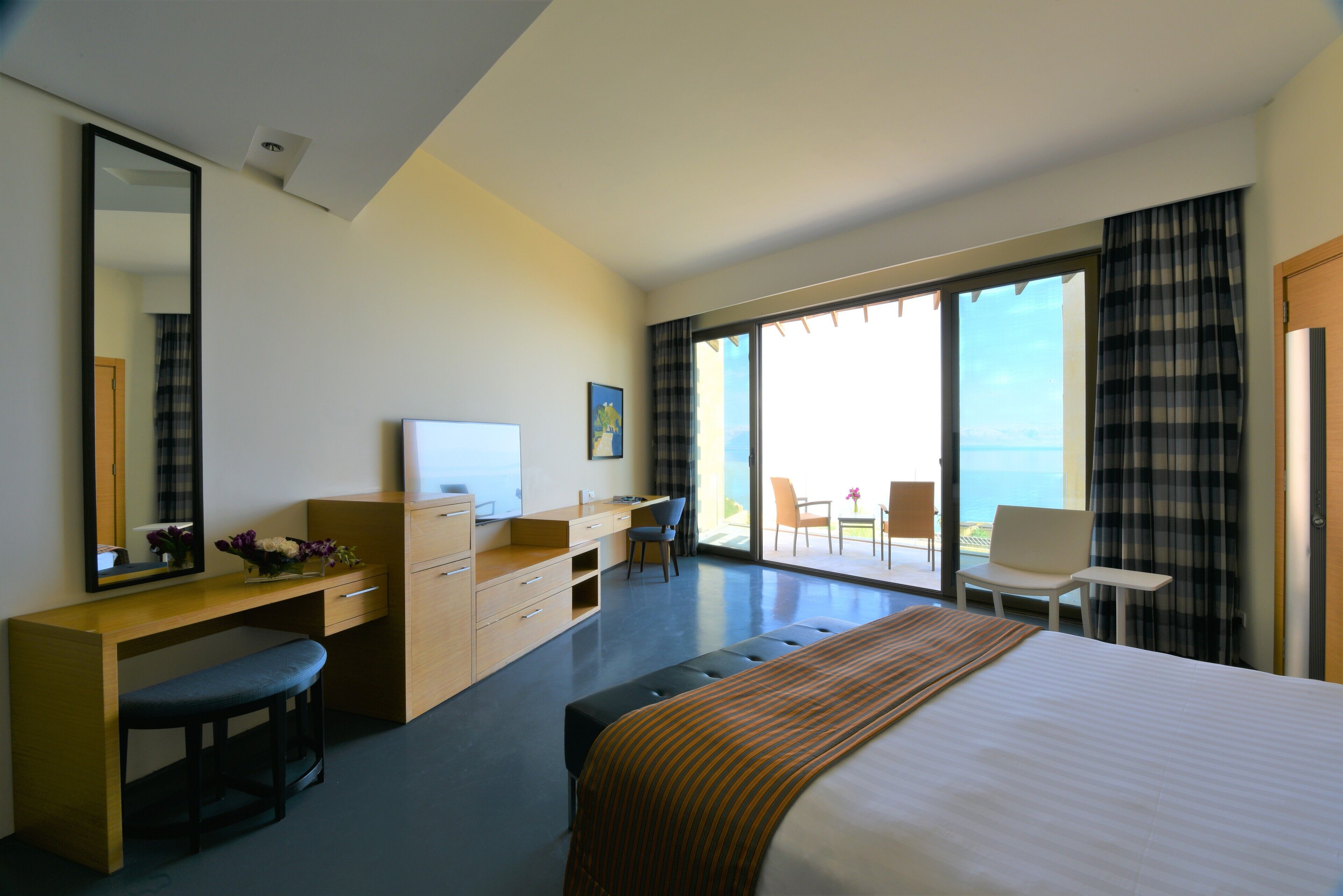 A modern hotel room with a large bed, a desk with a computer, a chair, and a view of the ocean through large windows.