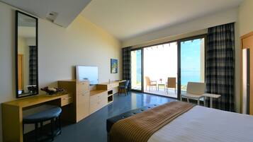 A modern hotel room with a large bed, a desk with a computer, a chair, and a view of the ocean through large windows.