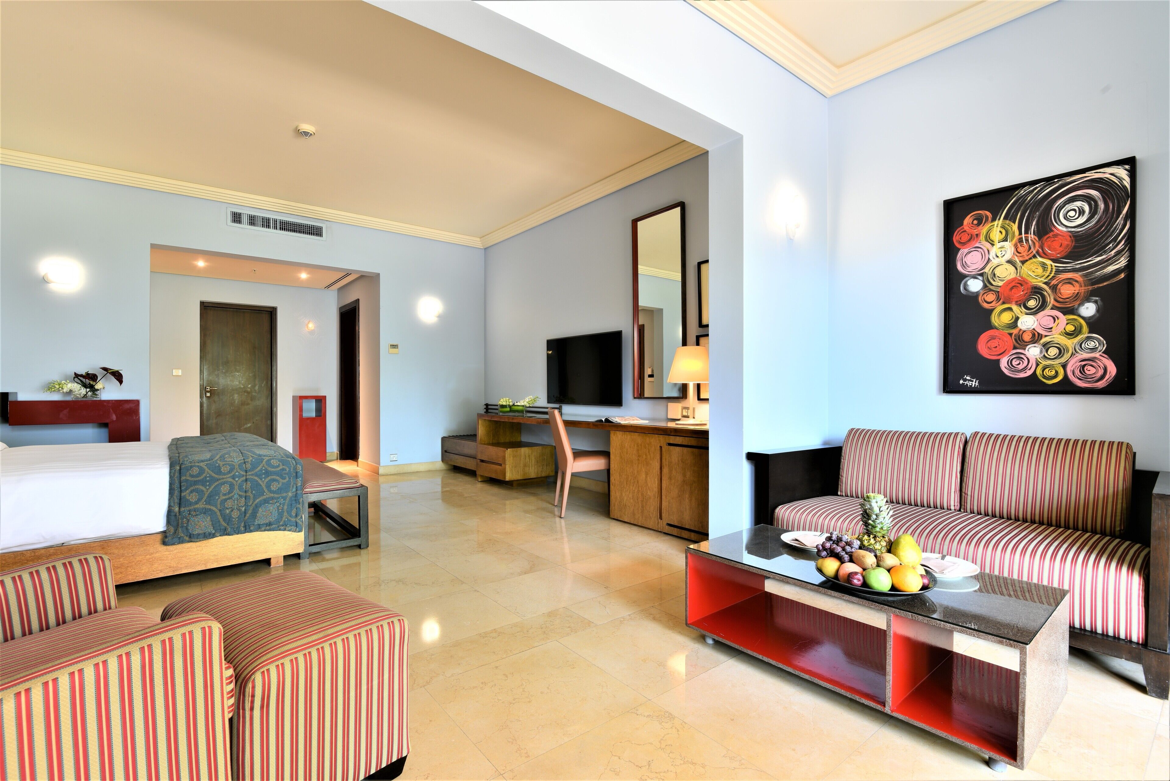 A hotel room with a bed, a sofa, a desk with a computer, a TV, and a painting on the wall.