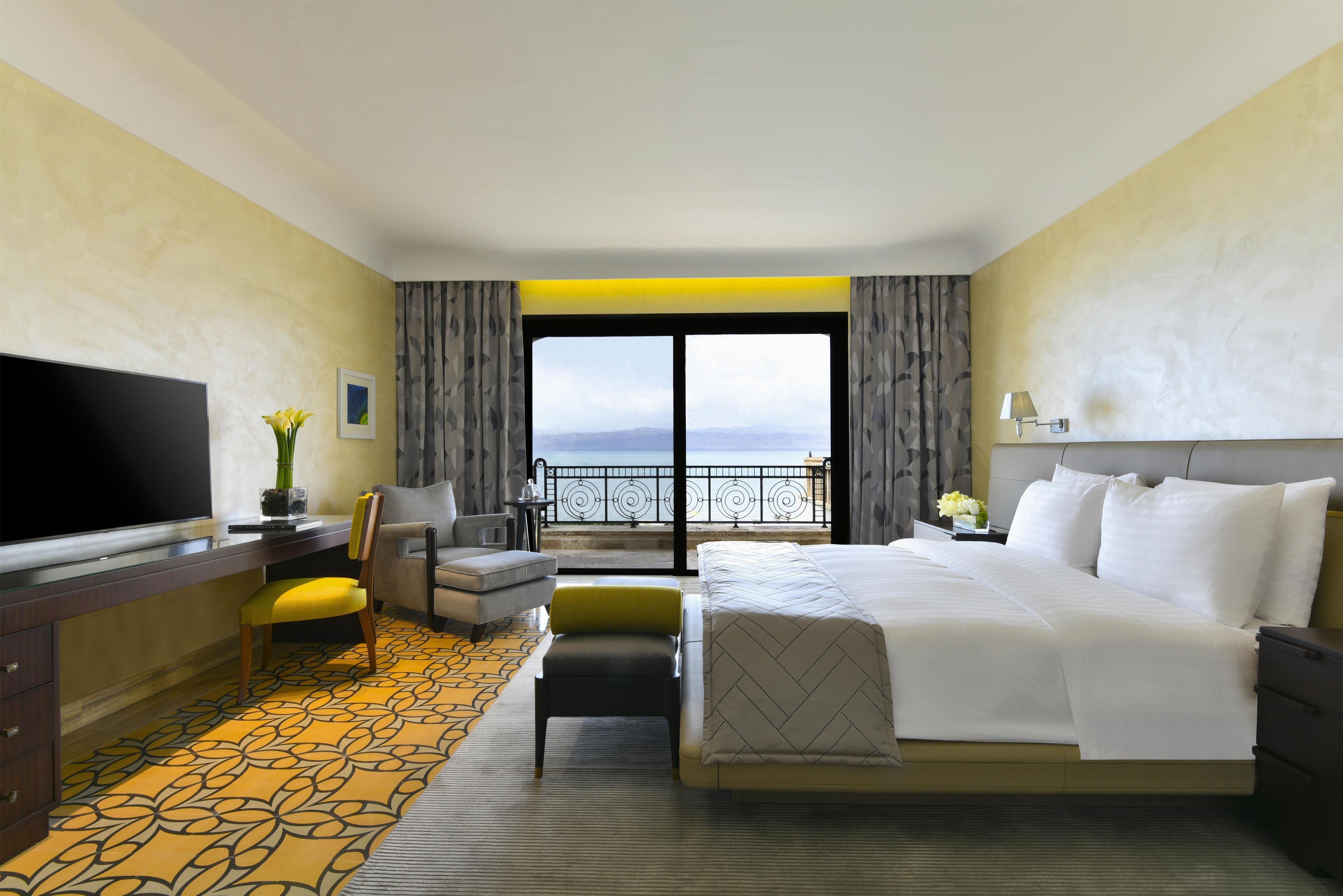 A hotel room with a bed, a TV, a desk, a chair, and a view of the sea.