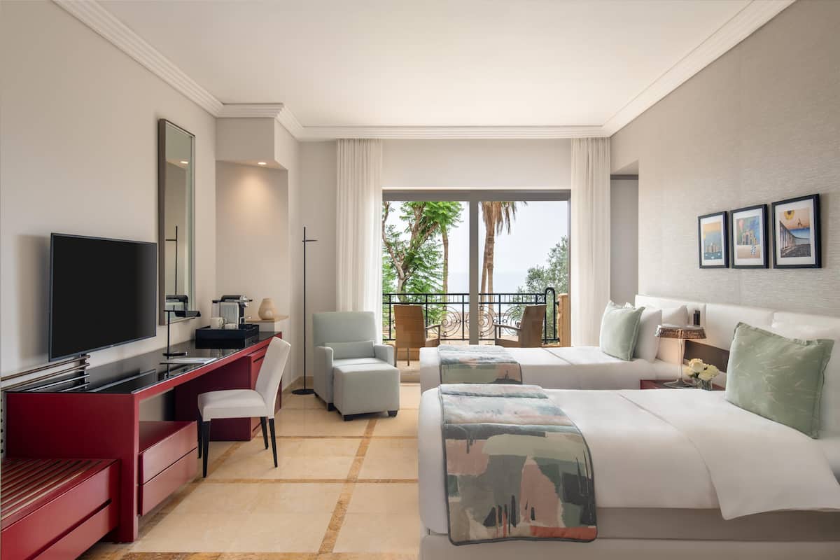 A hotel room with a TV, desk, two beds, a balcony with a view of palm trees, and framed pictures on the wall.