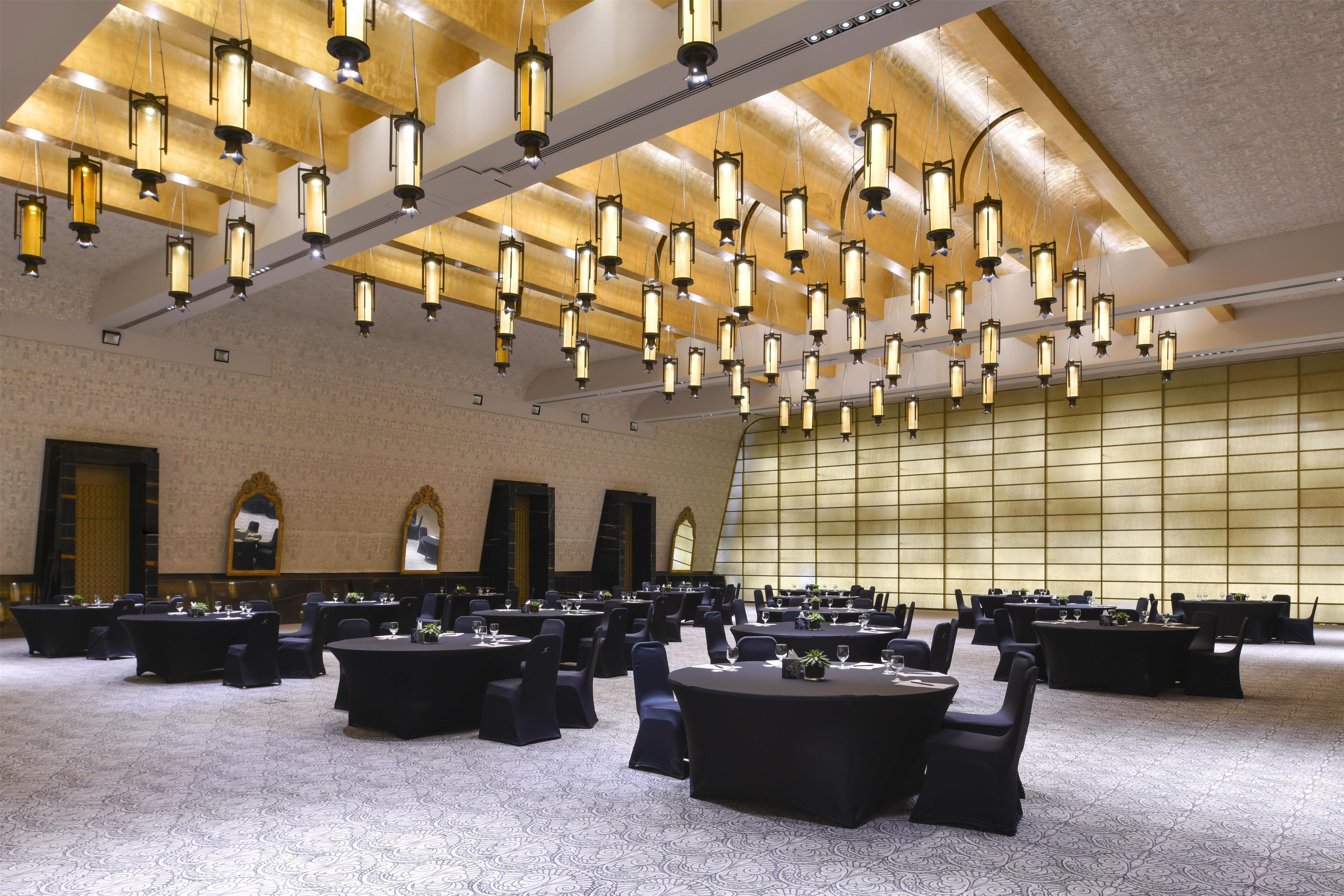 Un salón de conferencias con varias mesas y sillas, techo alto con lámparas colgantes y una pared grande con diseño de cuadrícula.