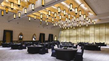 Un salón de conferencias con varias mesas y sillas, techo alto con lámparas colgantes y una pared grande con diseño de cuadrícula.