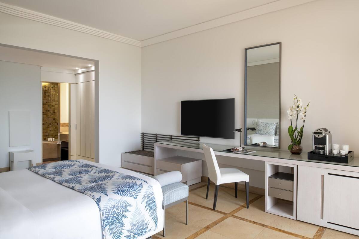 A modern hotel room with a bed, a desk with a flat-screen TV, a chair, and a large mirror.