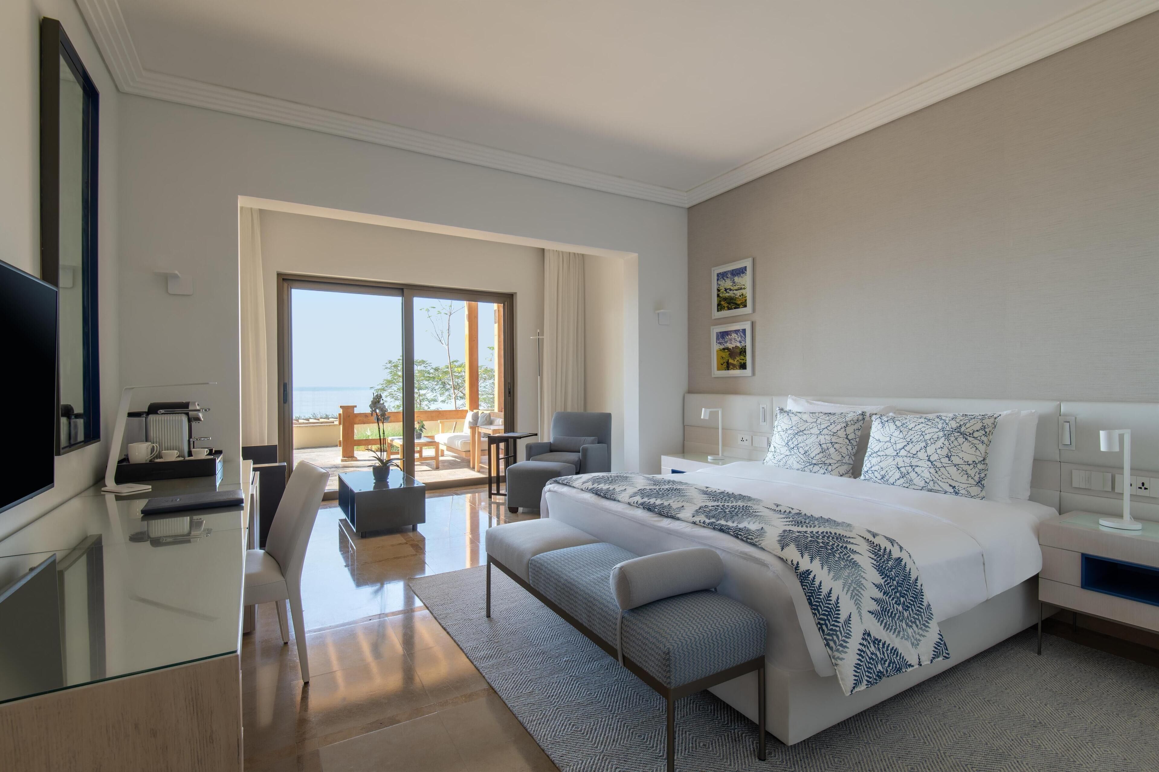 A modern hotel room with a large bed, a desk, a TV, and a balcony with a view.