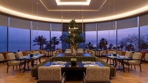 Un comedor espacioso con una mesa central, rodeado de sillas tapizadas, y ventanas amplias que ofrecen vista a palmeras y al océano.