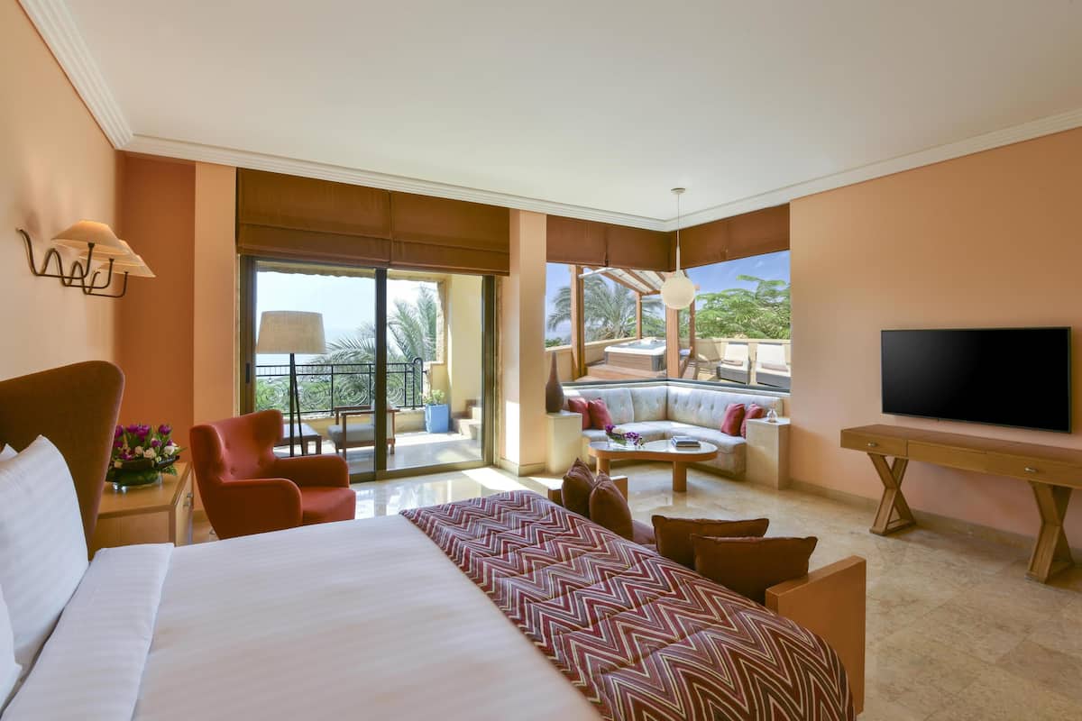 A hotel room with a large bed, a TV, a sofa, and a balcony with a view of palm trees.