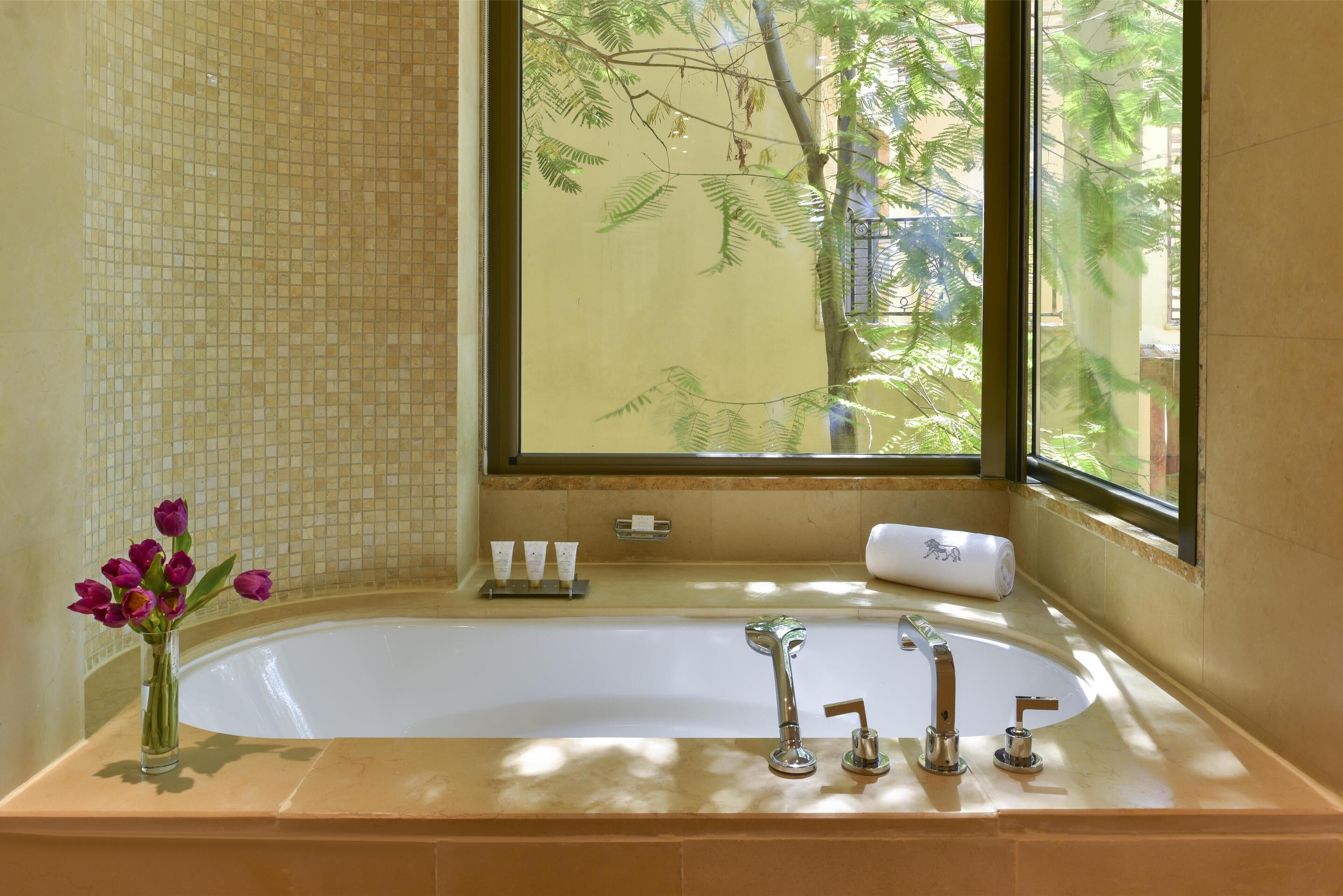 A bathroom with a large bathtub, a vase with flowers, and a window with a view of trees.