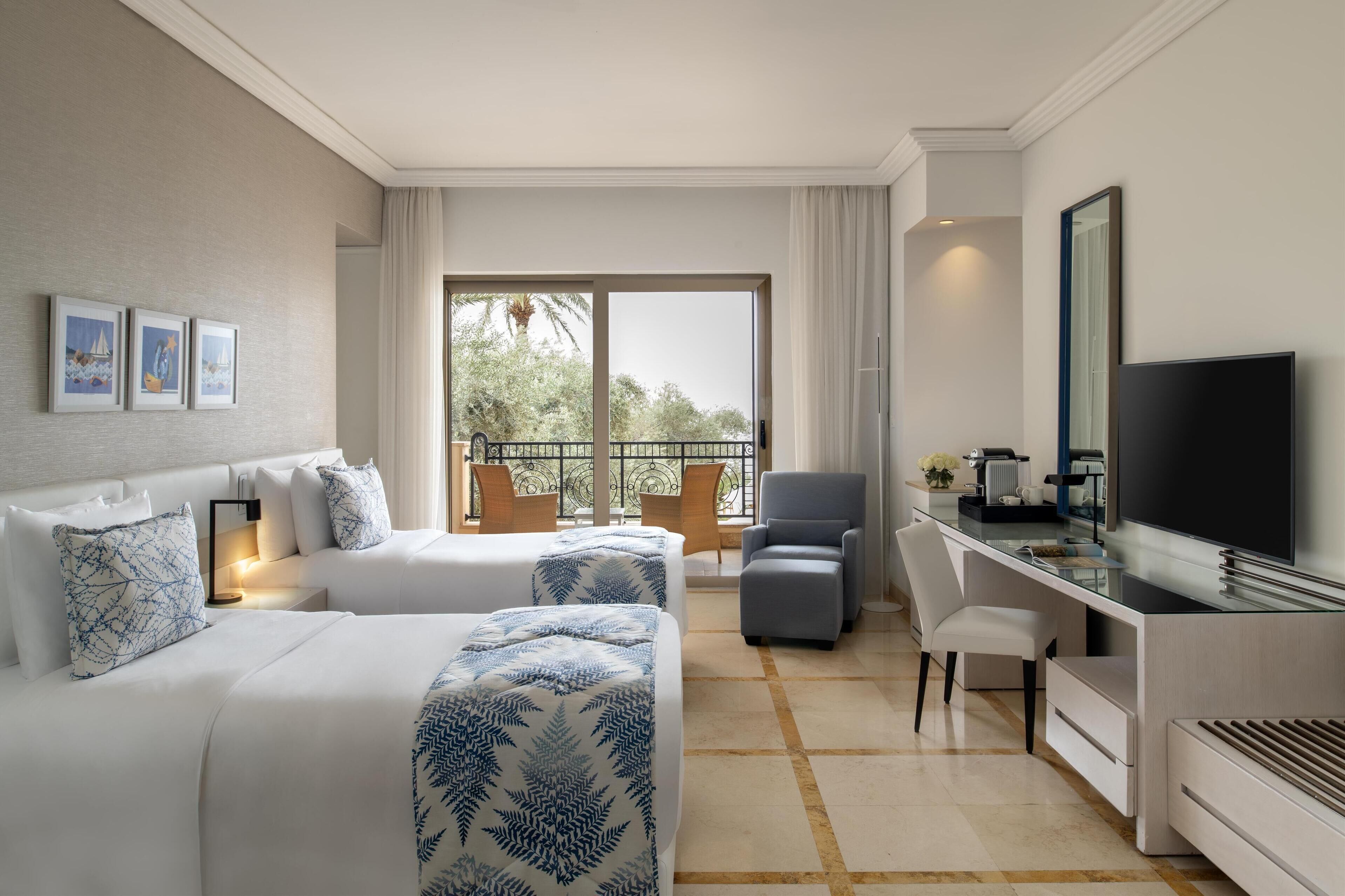 A hotel room with two beds, a desk, a TV, and a balcony with a view of trees.