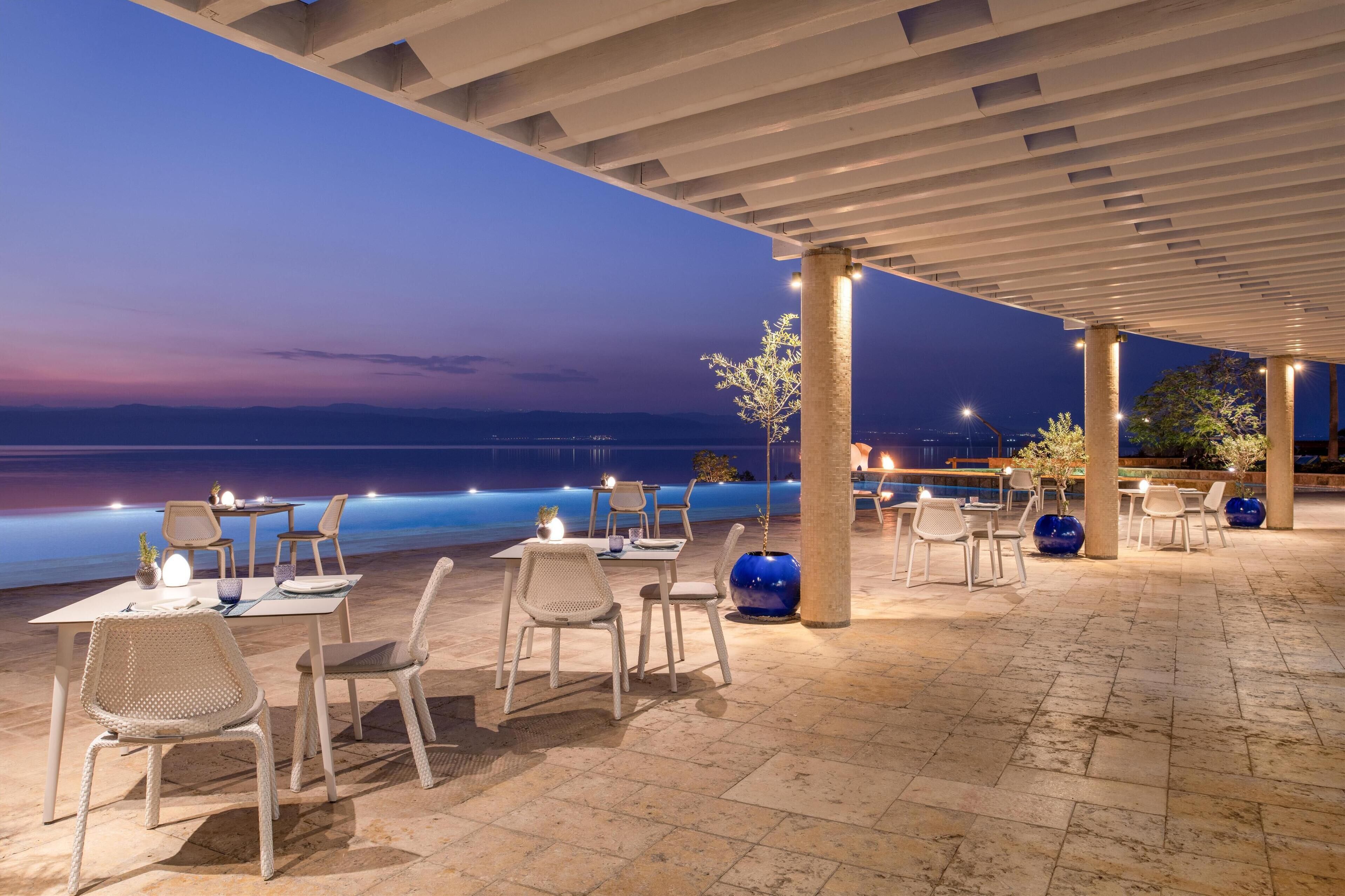 Un espace de restauration en plein air, avec des tables et des chaises, une pergola au-dessus et une vue sur la mer au crépuscule.