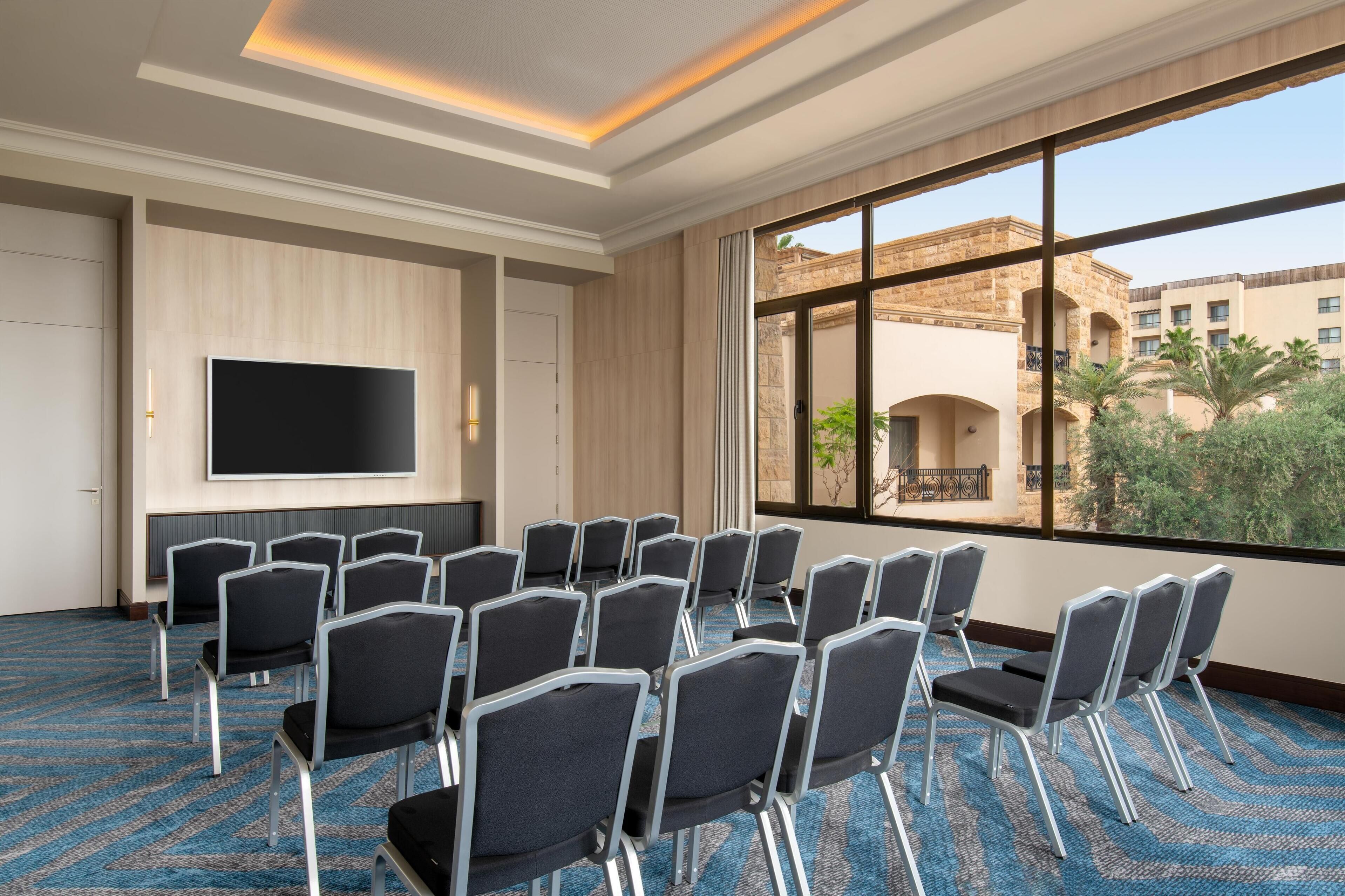 A conference room with a large TV screen, rows of chairs, and a view of a building with arches and palm trees.