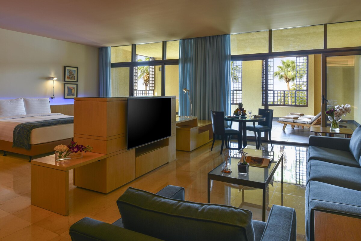 A modern hotel room with a bed, a TV, a sofa, a coffee table, and a dining area.