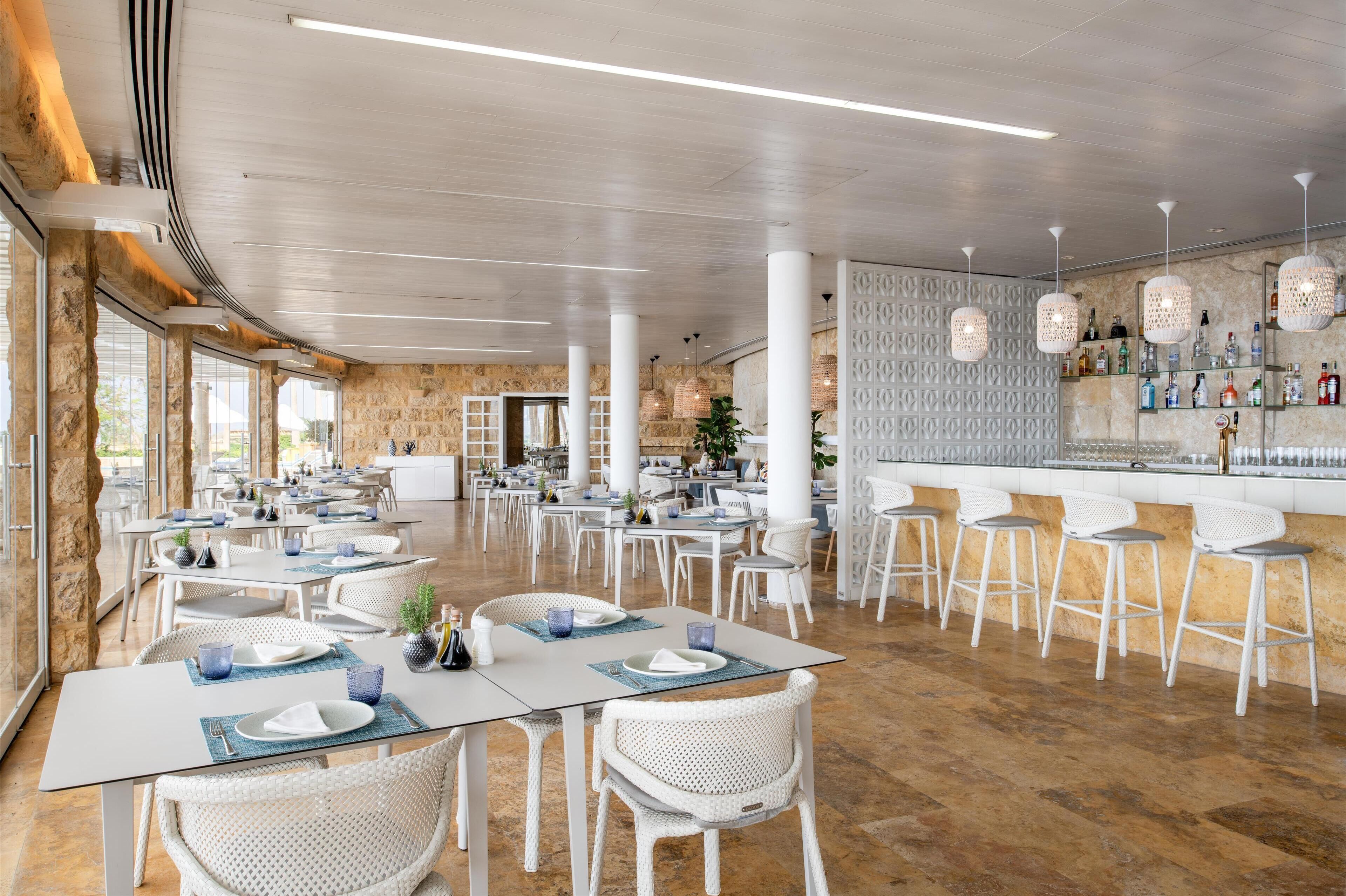 A spacious restaurant with white tables and chairs, a bar area with shelves of bottles, and a stone wall with a geometric pattern.