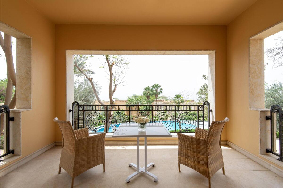 A balcony with a table and two wicker chairs, overlooking a swimming pool and greenery.