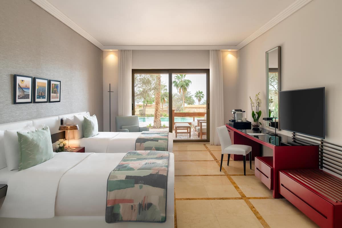 A hotel room with two beds, a desk with a TV, and a balcony with a view of palm trees.