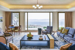 A spacious living room with a large window offering a view of the sea, a dining area with a table set for two, and a comfortable seating arrangement. - Kempinski Hotel Ishtar Dead Sea (Sweimeh)